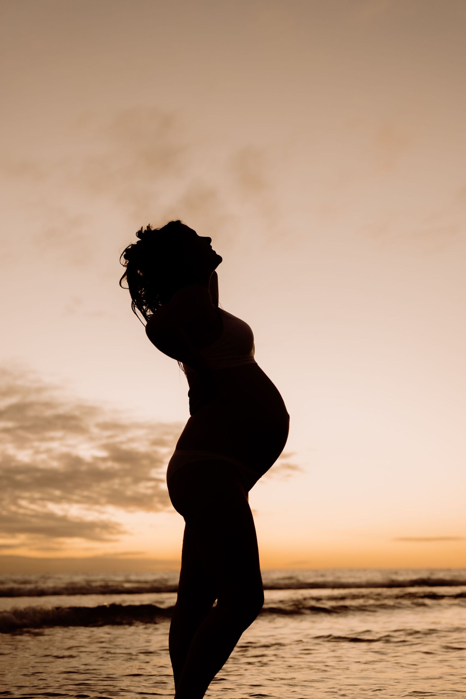 Silhouette of a pregnant woman standing on a beach at sunset, with ocean waves and golden sky in the background.