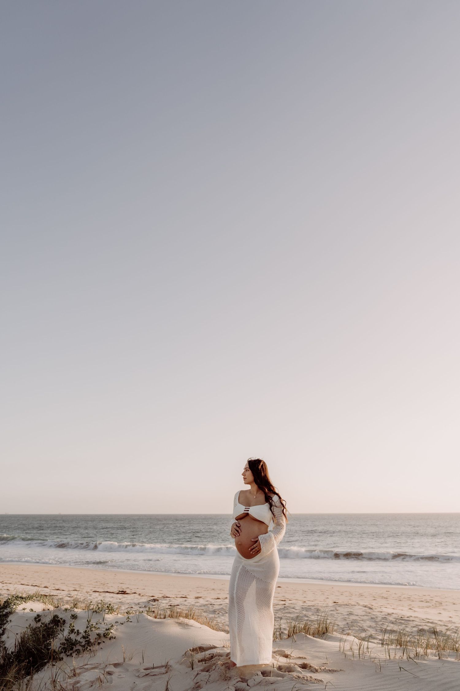A pregnant woman in white dress stands on a sandy beach at sunset with ocean waves in the background.