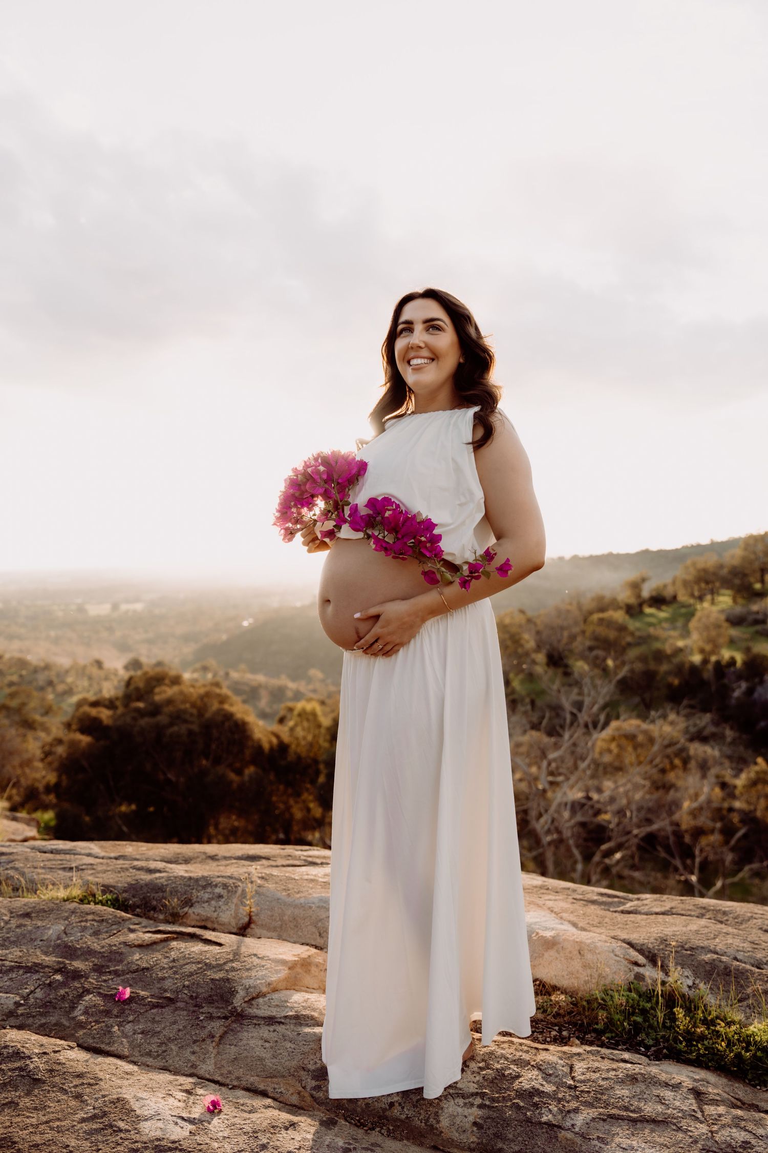 Pregnant woman in white dress holding pink flowers on rocky overlook with scenic landscape backdrop.