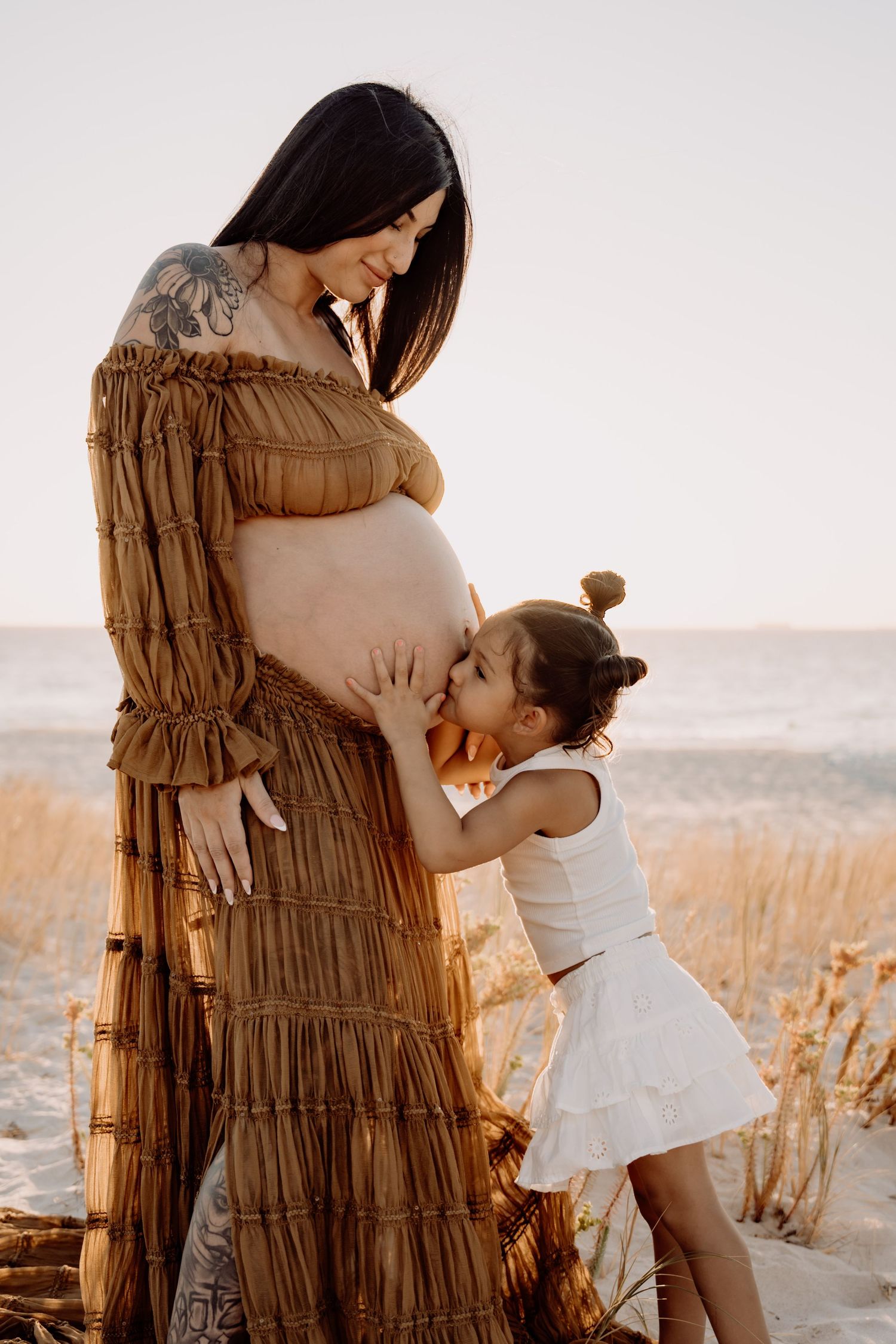 Pregnant woman in brown dress with young daughter kissing her belly on a sandy beach at golden hour.