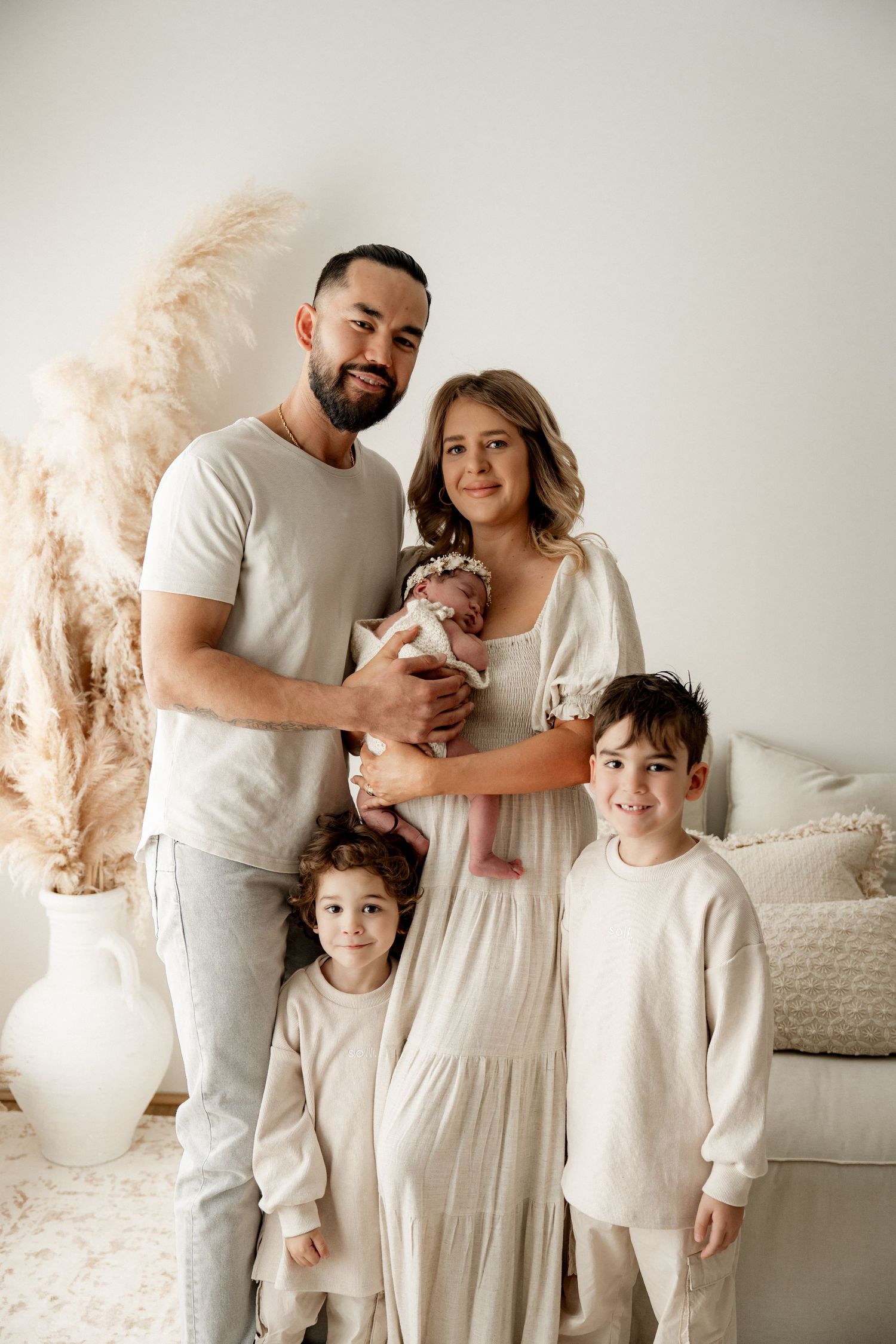 Family of five posing together in neutral tones, parents holding newborn baby with two young children smiling in bright studio.