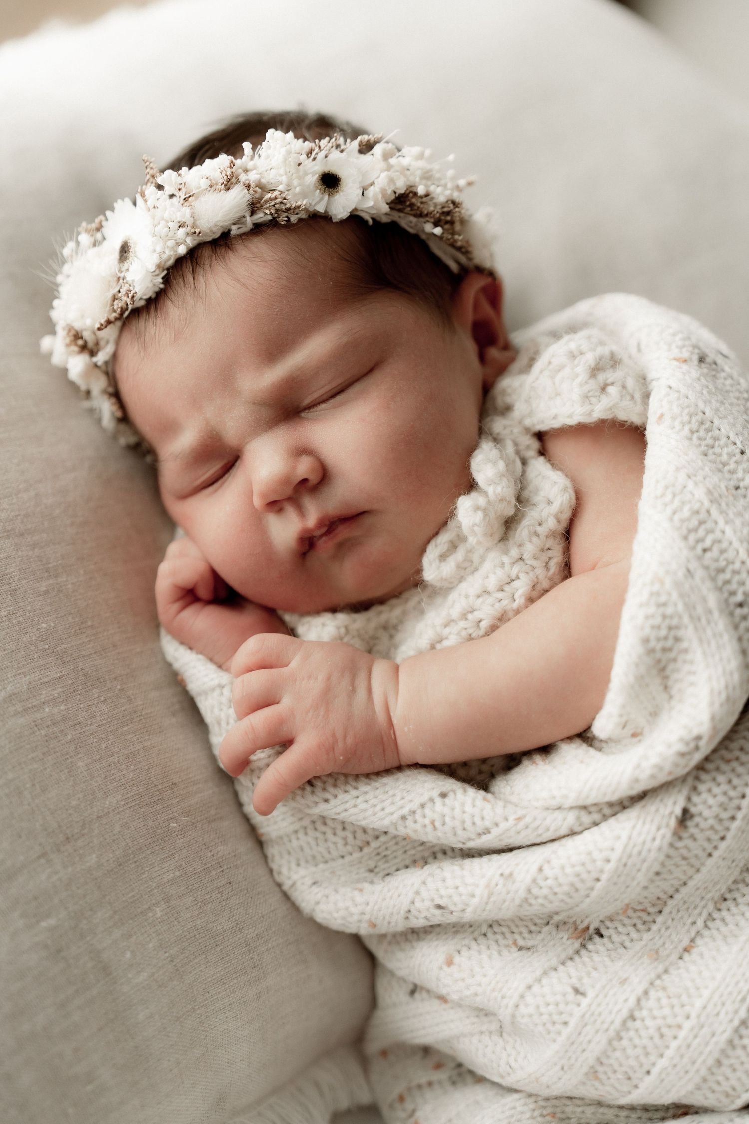 Sleeping newborn baby girl wrapped in white knit blanket wearing a delicate floral crown headband.