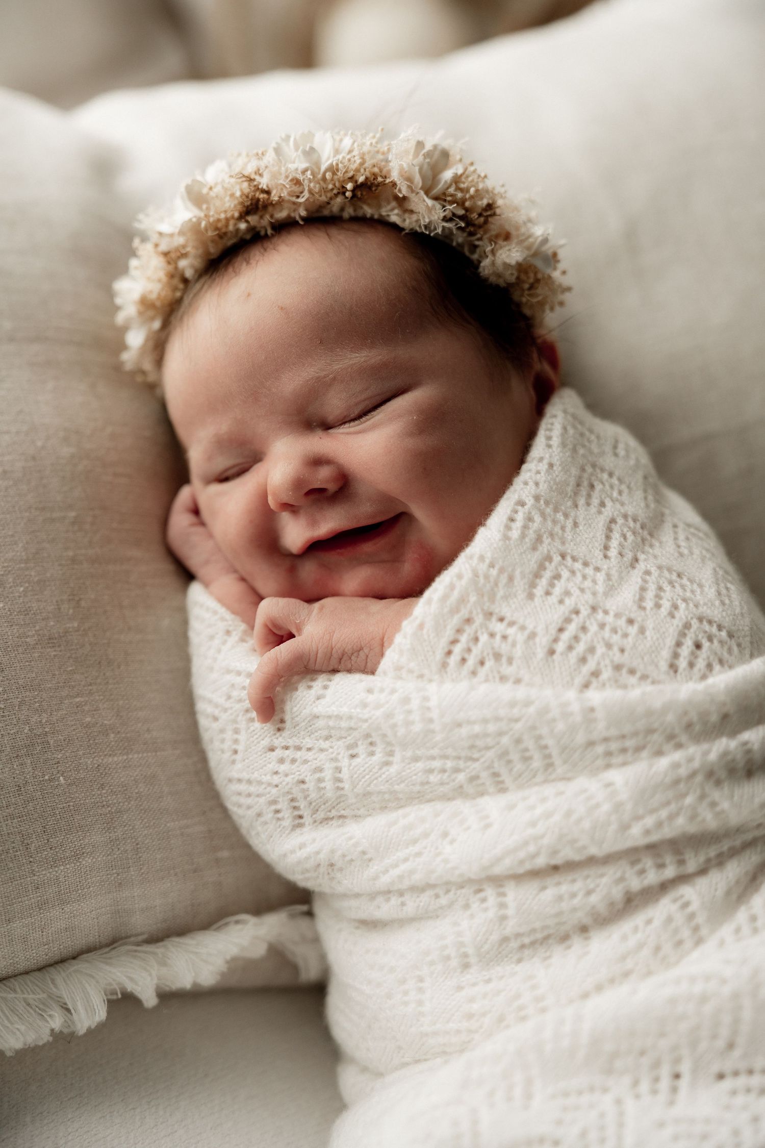 Smiling newborn baby wrapped in white knit swaddle blanket wearing a delicate floral crown, sleeping peacefully.