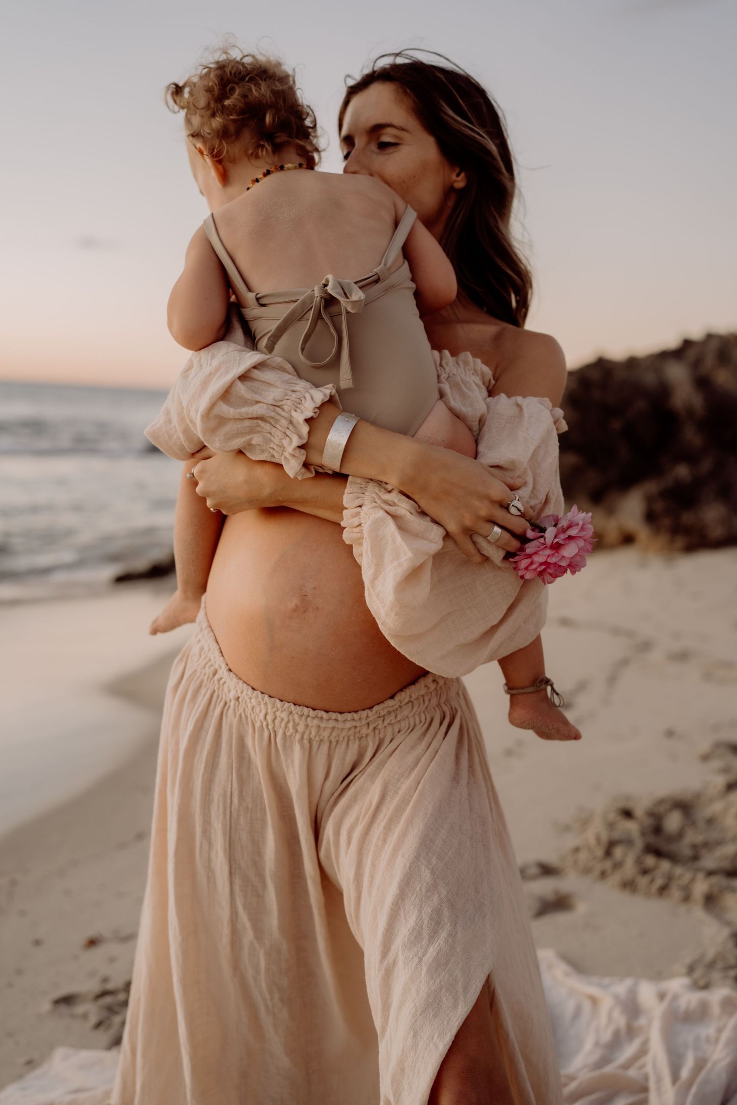 Maternity beach photo at sunset showing pregnant woman in flowy dress holding toddler.