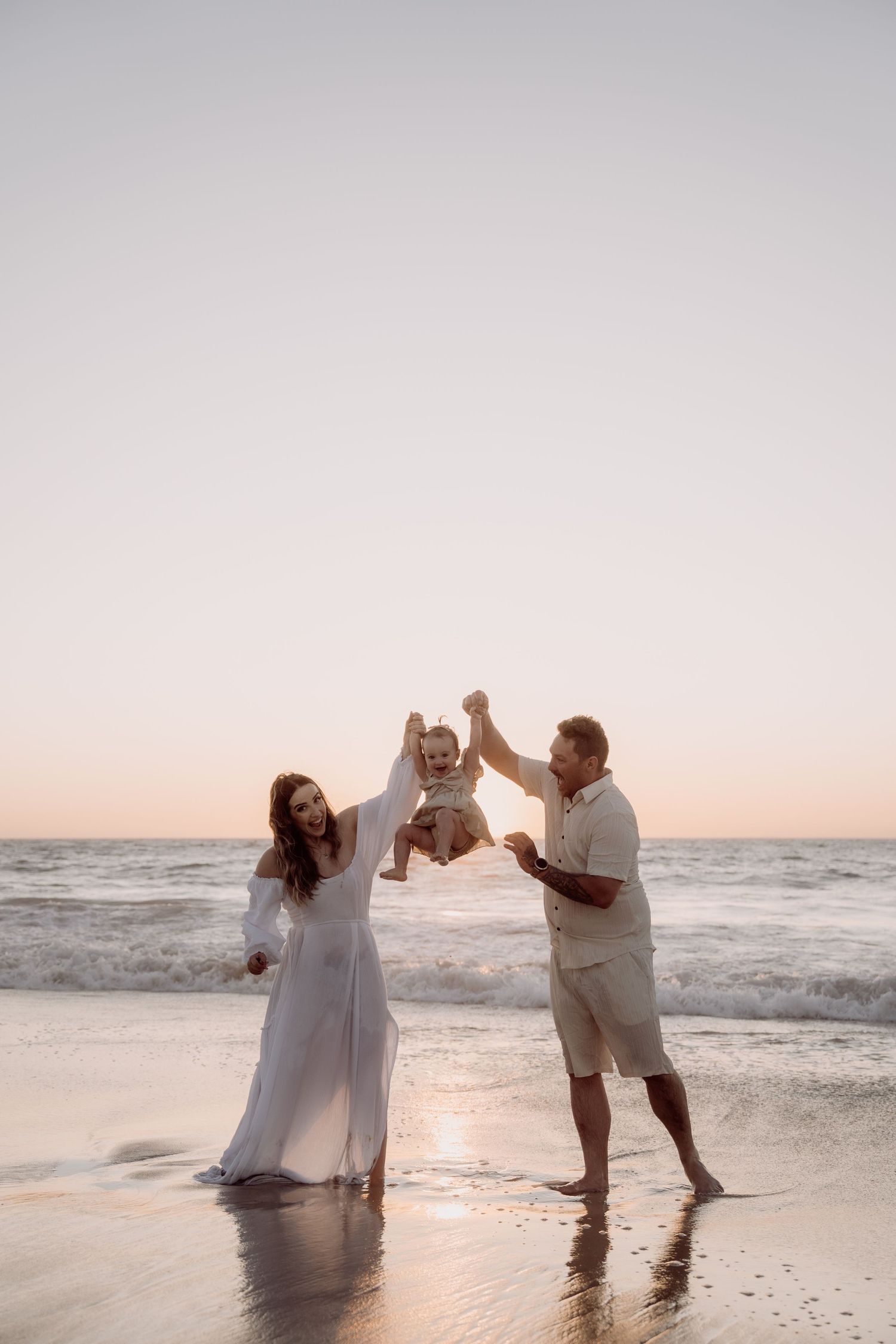 A family playing together at sunset on a beach shore with crashing waves and soft pink sky in the background.