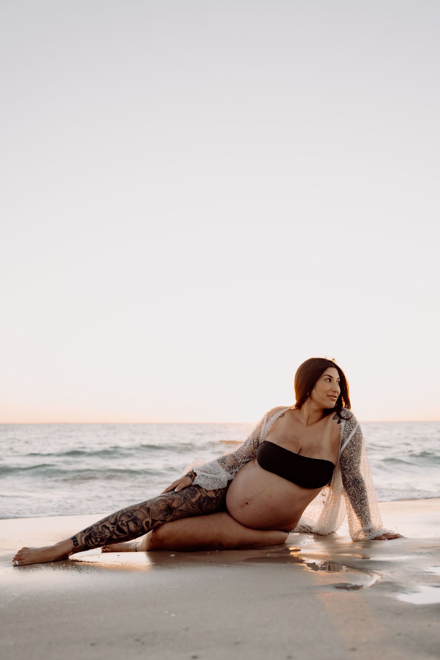 Pregnant woman with tattoos sitting on a beach in a black bikini top, wearing a sheer wrap, with ocean waves behind her.