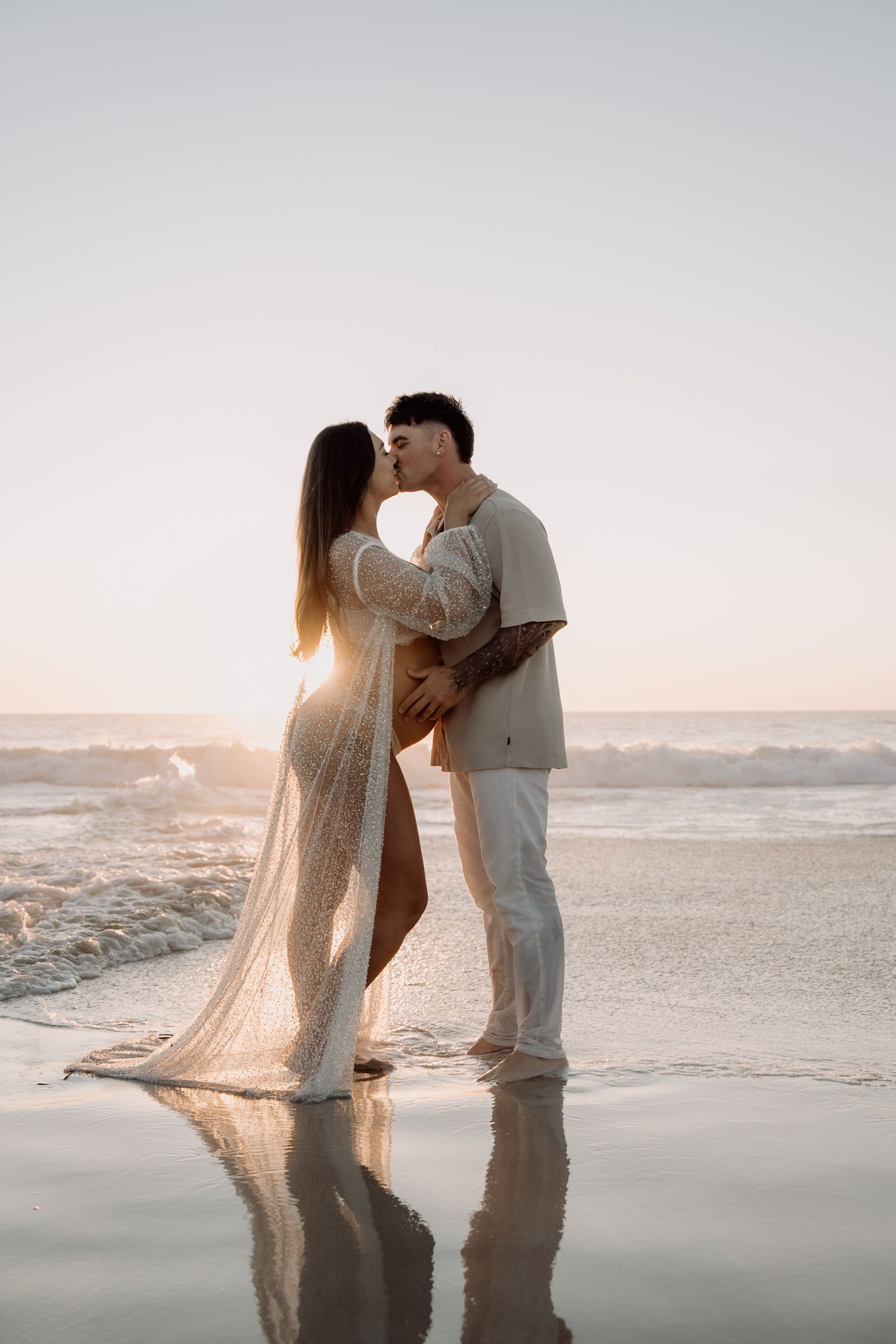 A romantic couple shares an intimate moment on a misty beach at sunset with flowing fabric creating an ethereal scene.