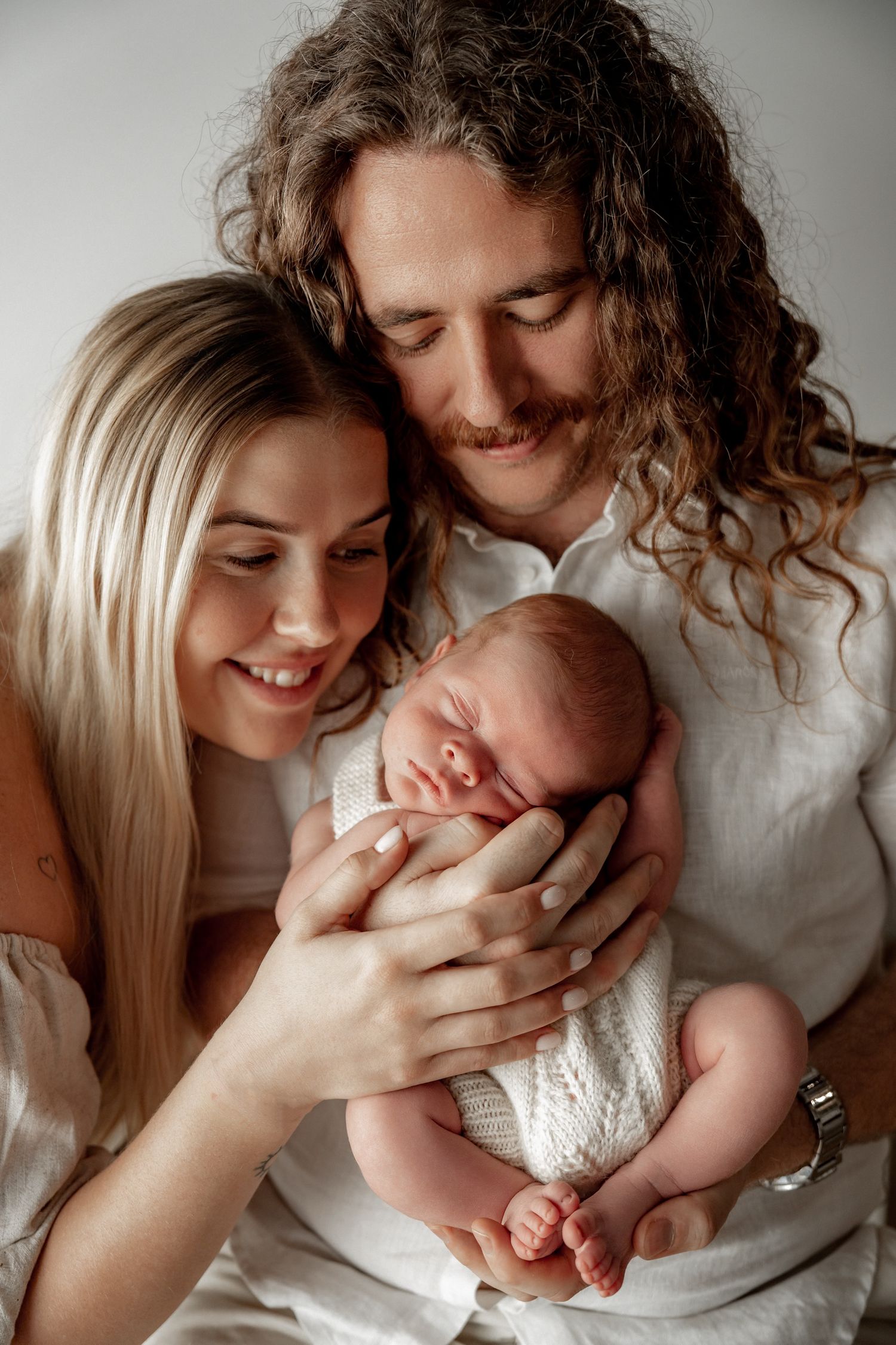 A young couple with long hair tenderly holds their sleeping newborn baby, dressed in white, against a soft light background.