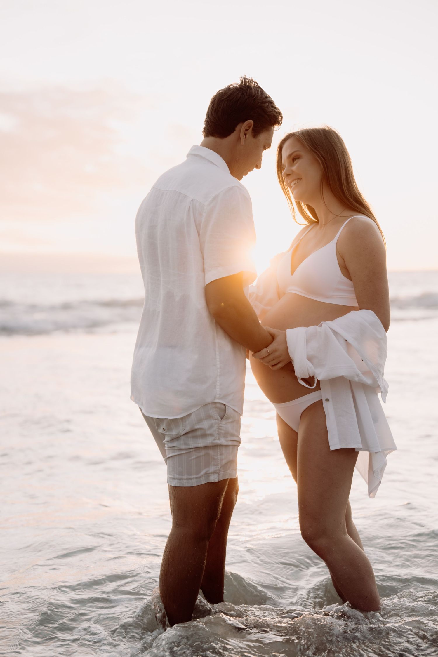 Couple standing in ocean waves at sunset, woman in white bikini with open shirt, man in white shirt and shorts.