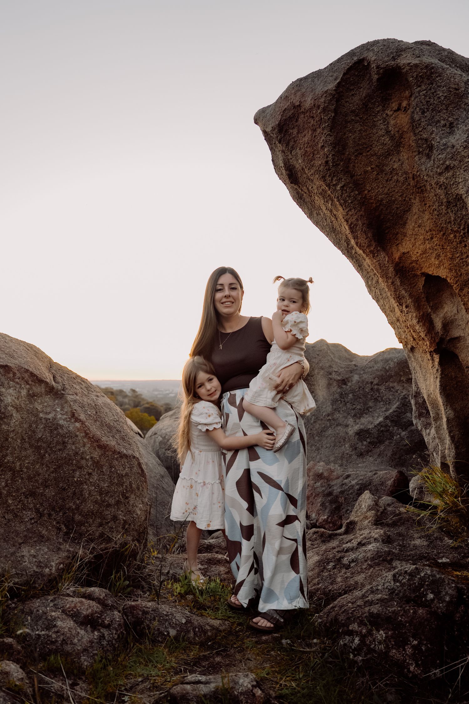 A figure holds a white cat while standing among large rock formations at sunset.