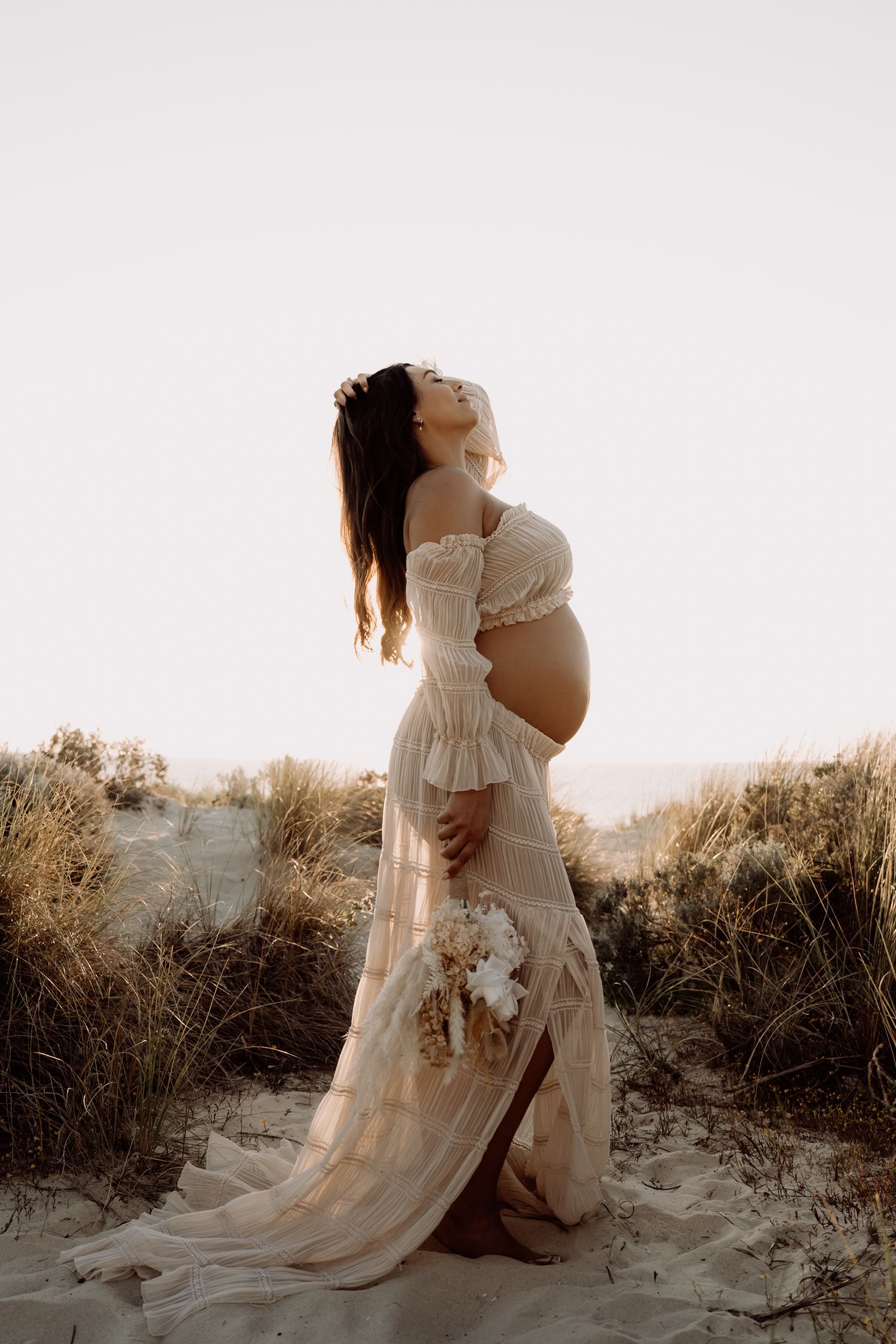 Maternity photoshoot at sunset shows silhouetted figures in flowing white dresses on a sandy beach with golden light.