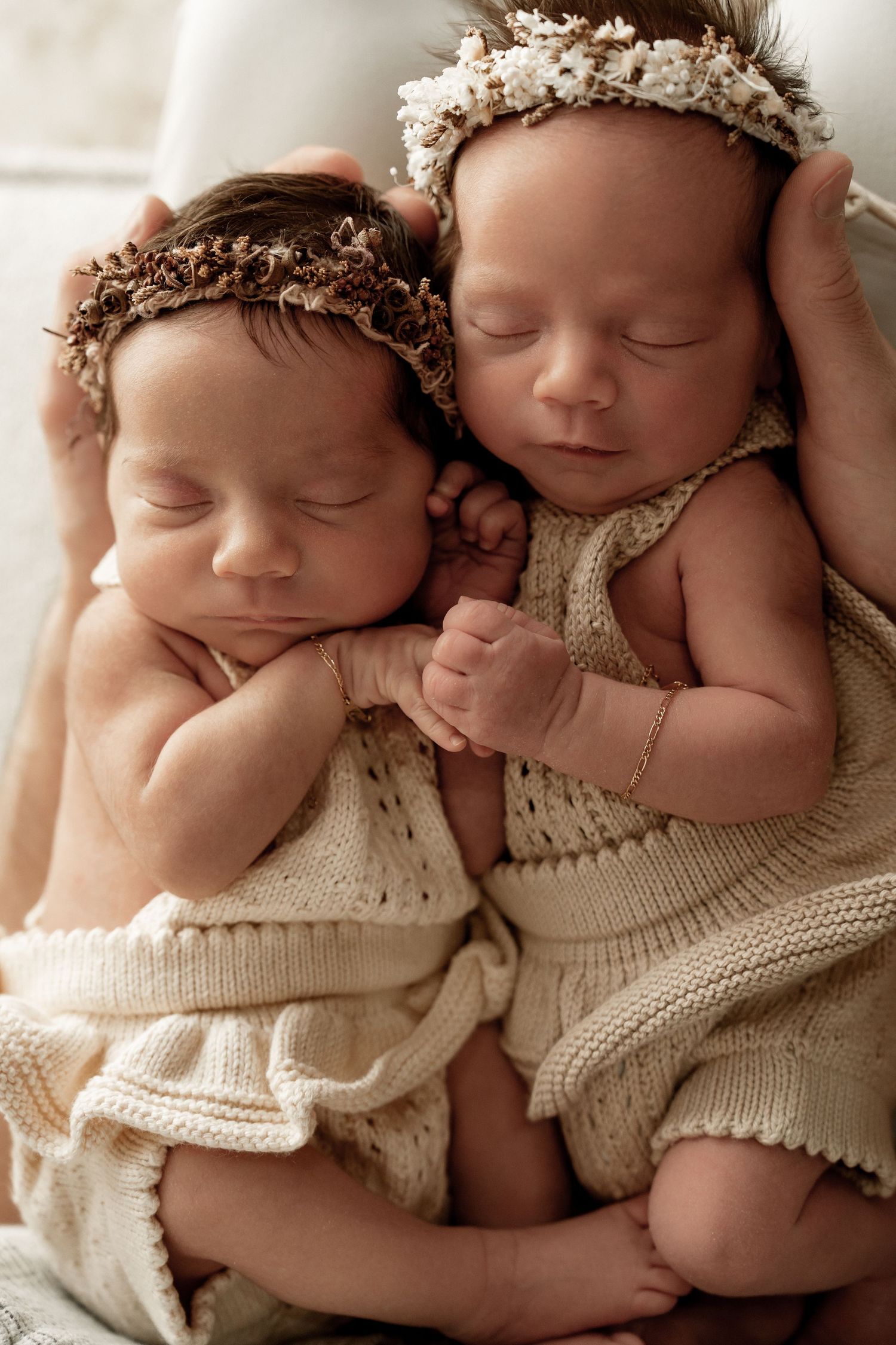 Two sleeping newborns in knit rompers wearing delicate floral headpieces rest peacefully on cream-colored fabric.