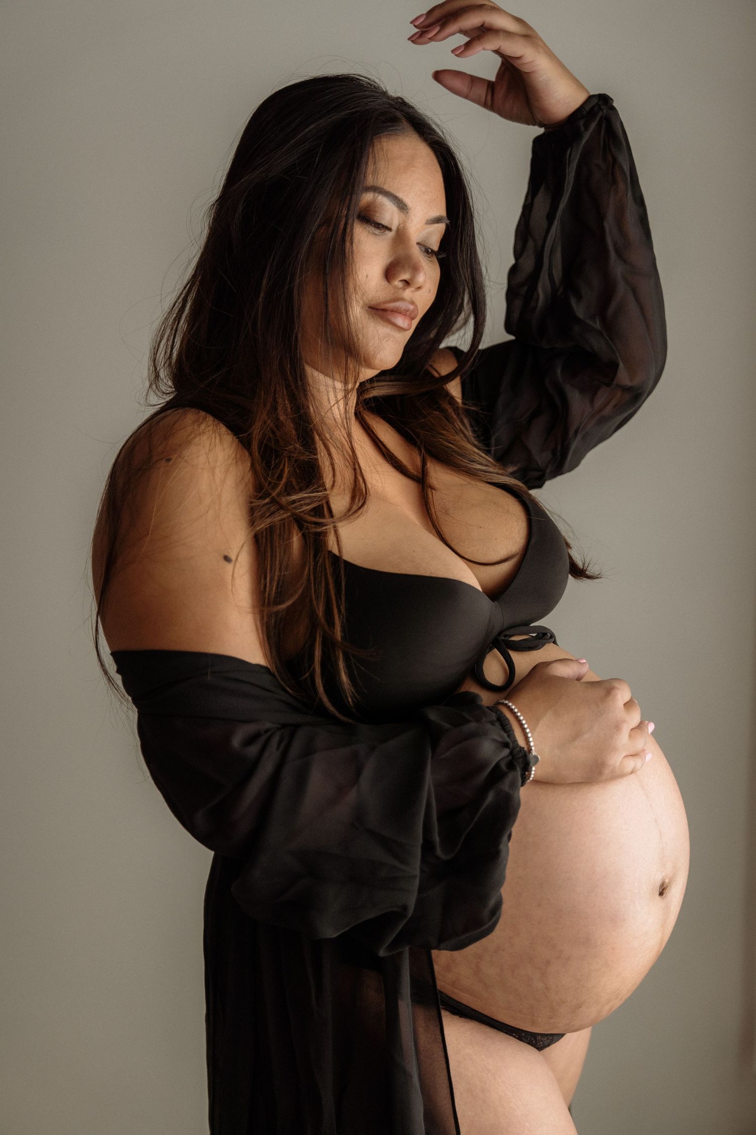Artistic maternity photo of an expectant mother in a black robe posing against neutral backdrop in soft lighting.