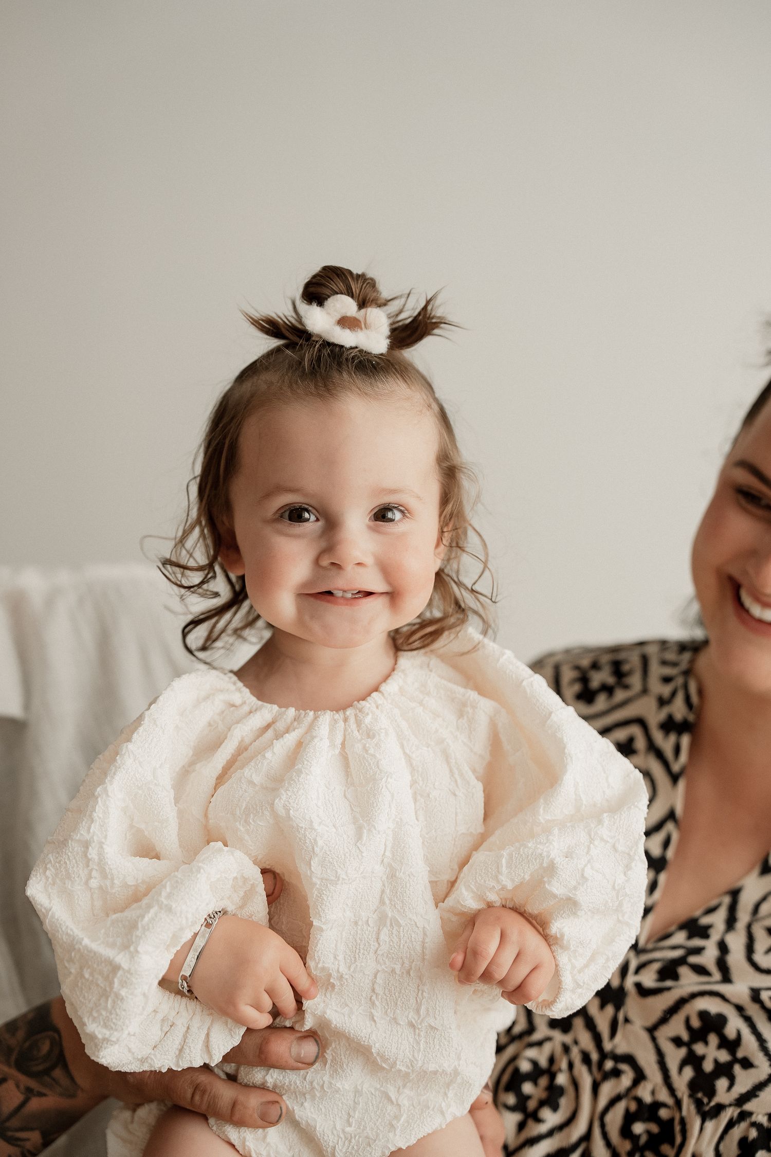 A baby wearing a delicate white lace dress smiles while sitting on a patterned blanket.