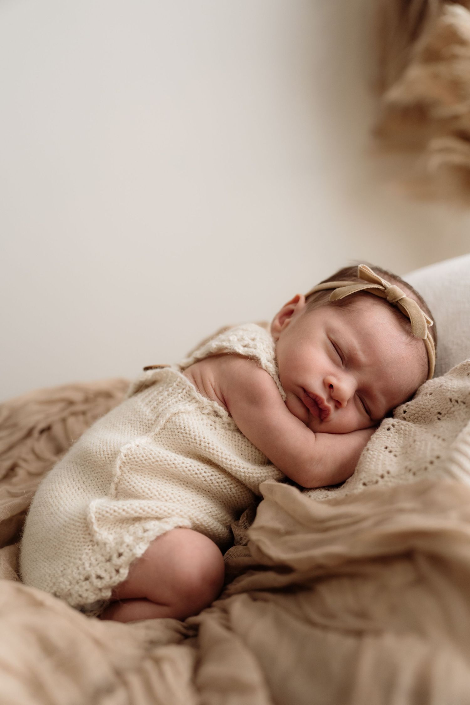 A sleeping baby wearing a white lace dress rests peacefully on beige bedding in soft natural lighting.