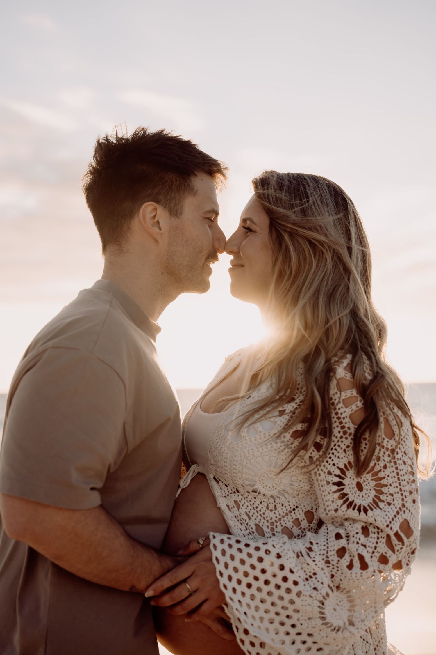 Couple sharing a romantic almost-kiss at golden hour sunset, woman in crochet dress with glowing backlight.