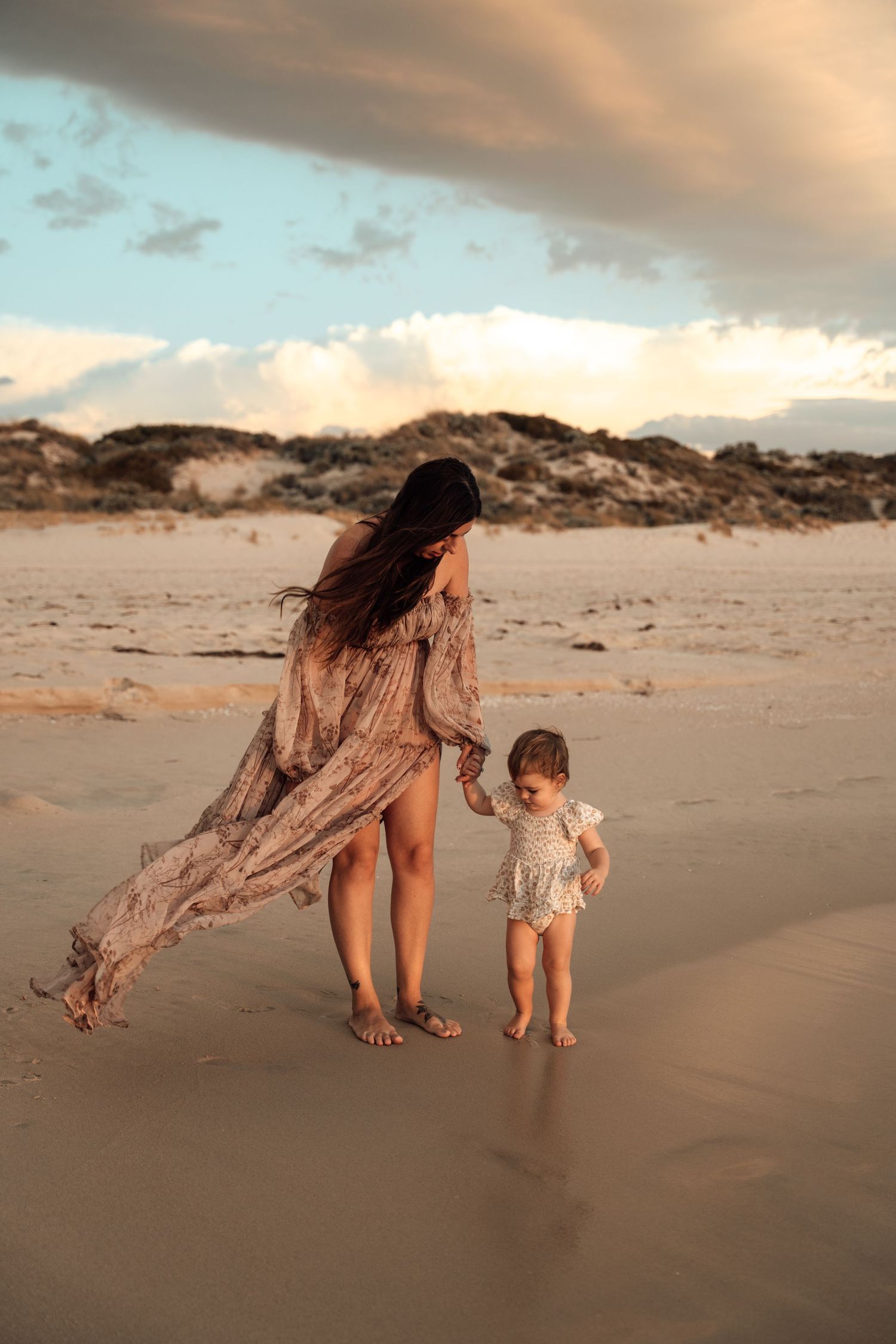 Flowing dress catches wind as mother and child walk along beach at sunset.
