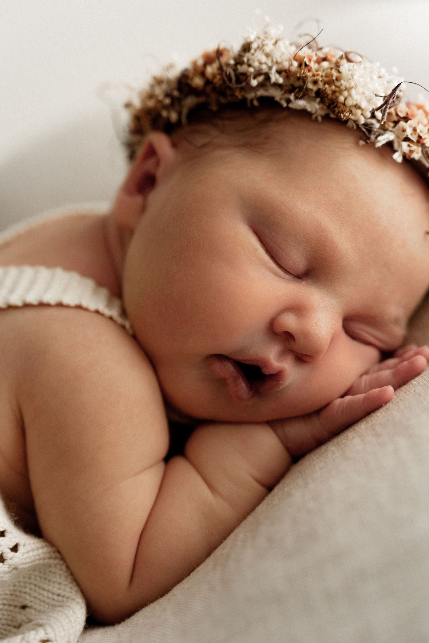 Sleeping baby wearing floral crown rests peacefully on soft white blanket.