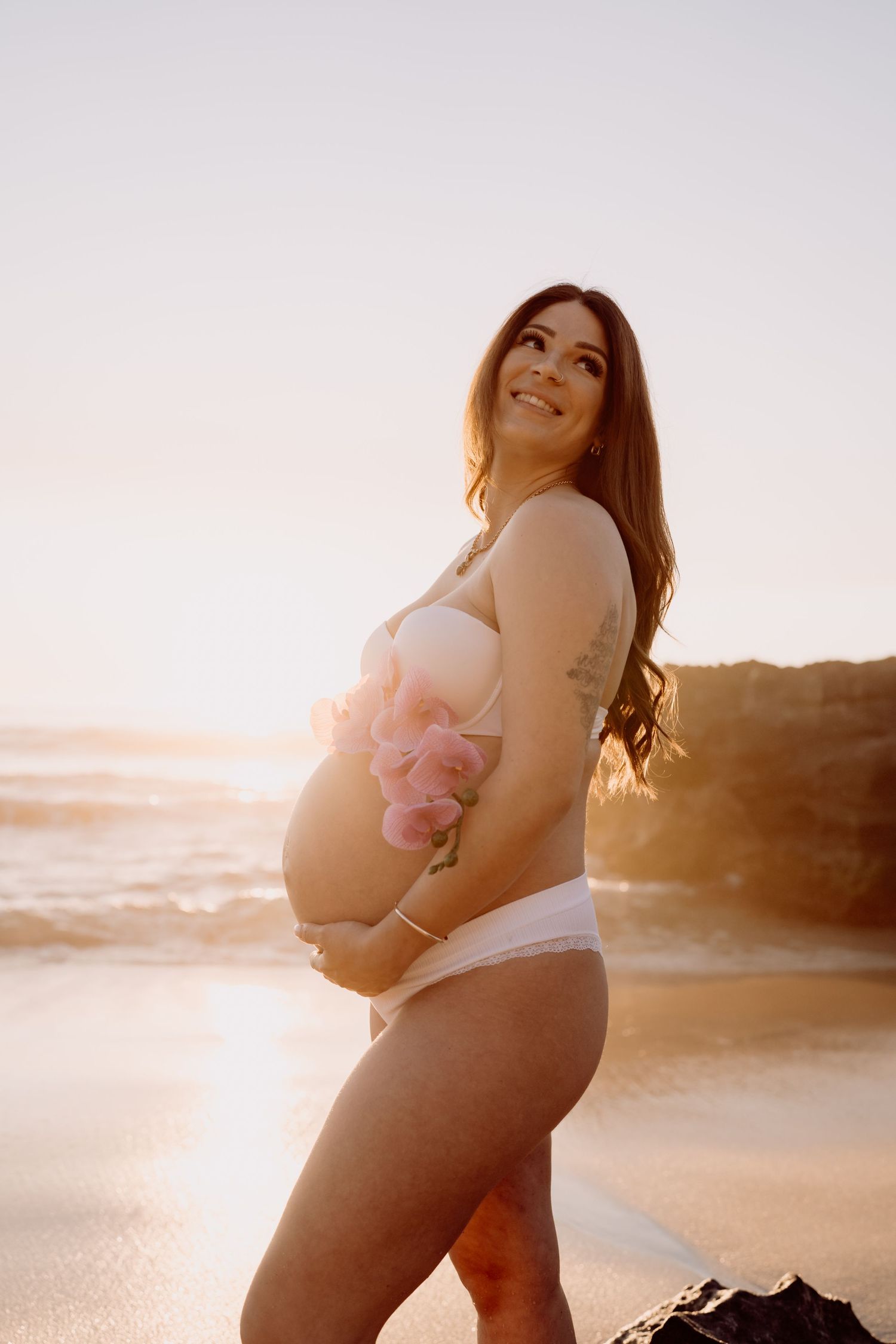 A pregnant woman in white lingerie poses on a beach at sunset with golden light creating a dreamy silhouette effect.