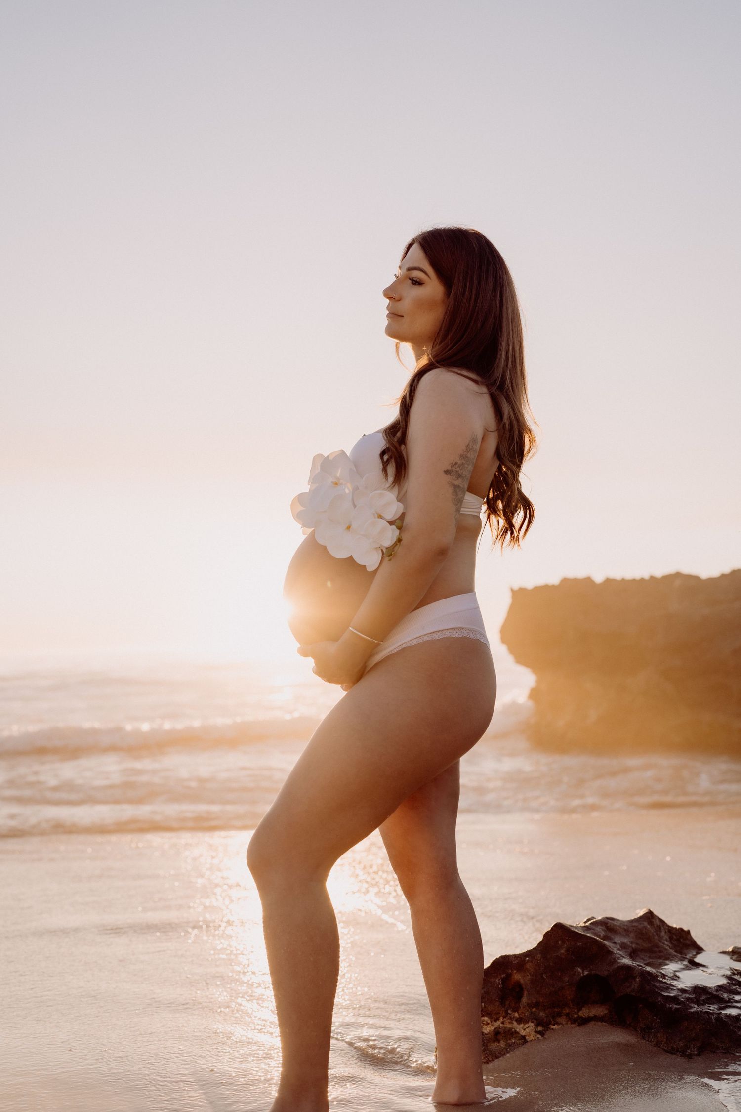 A pregnant woman in white swimwear poses on a beach at sunset with rocks and ocean waves in the background.