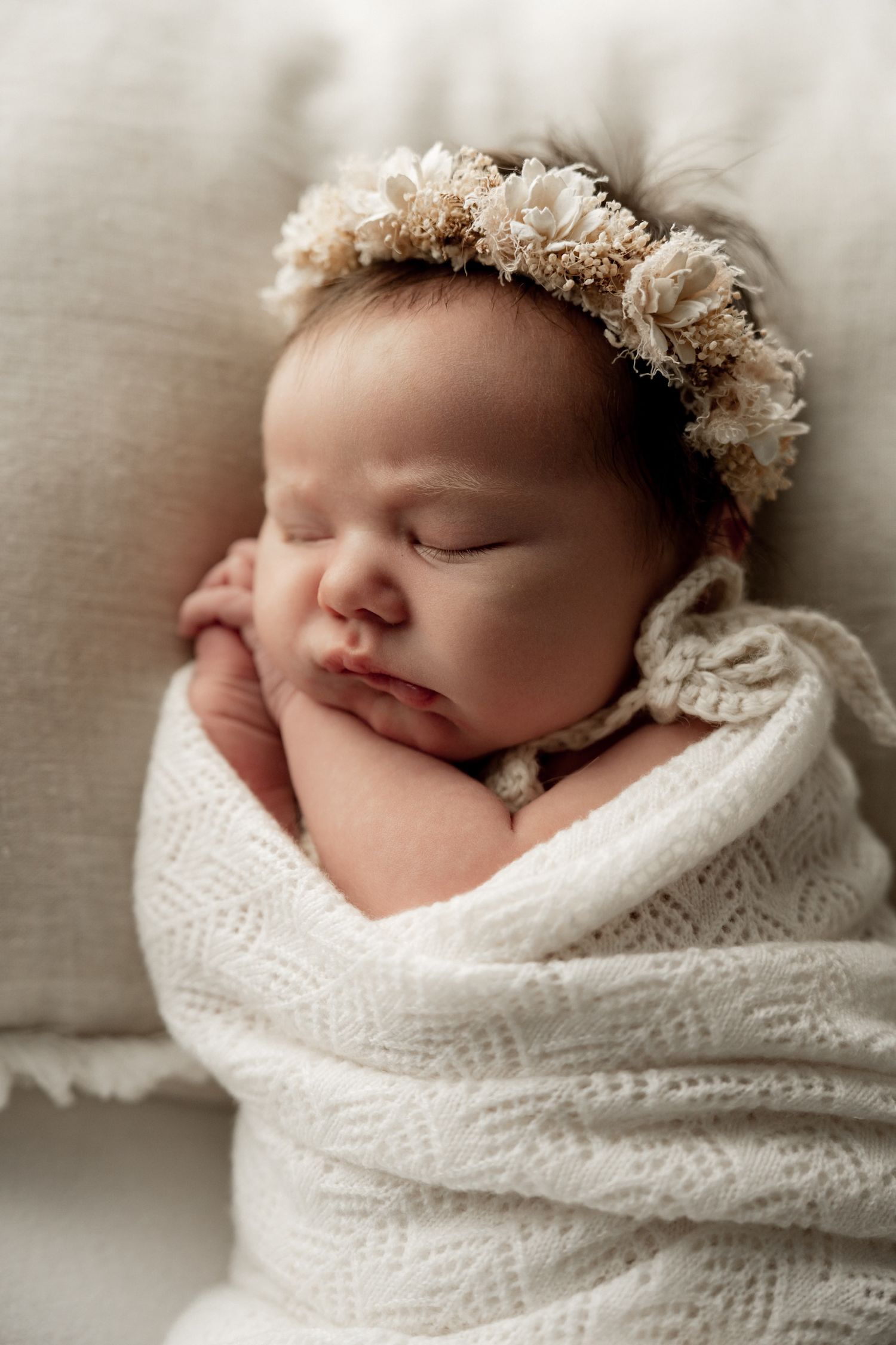 Sleeping newborn baby wrapped in white knit blanket wearing a delicate floral crown headband.