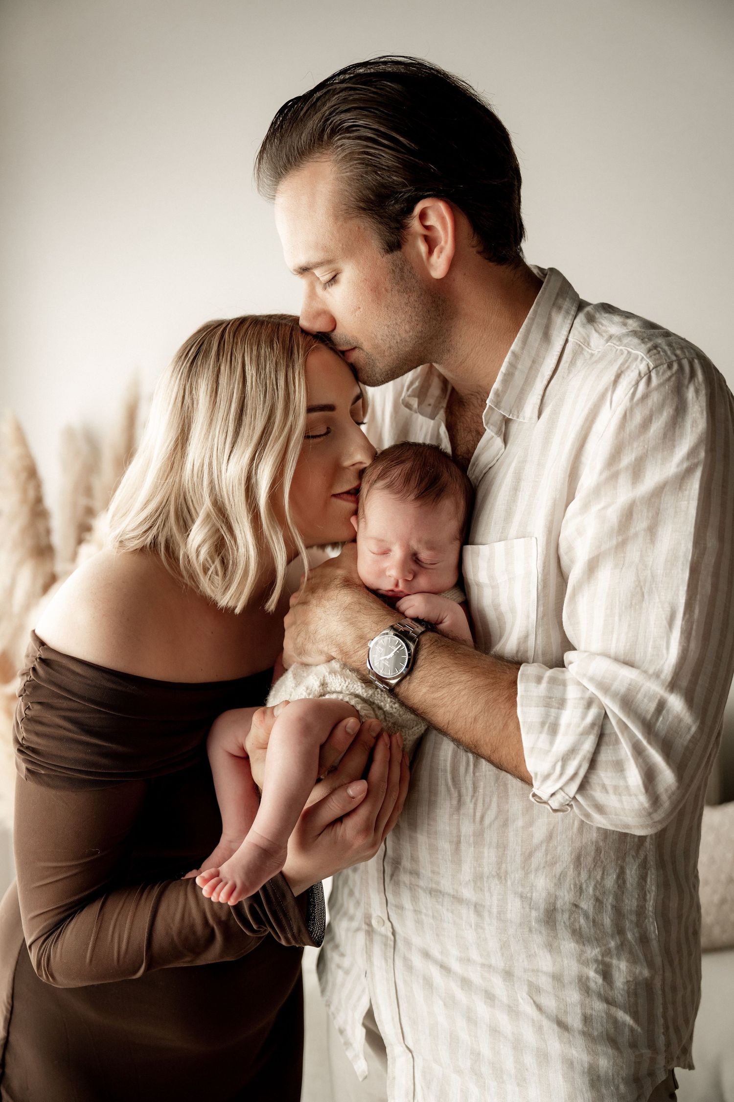 A couple tenderly holds and kisses their sleeping newborn baby in a soft, neutral-toned portrait photo.