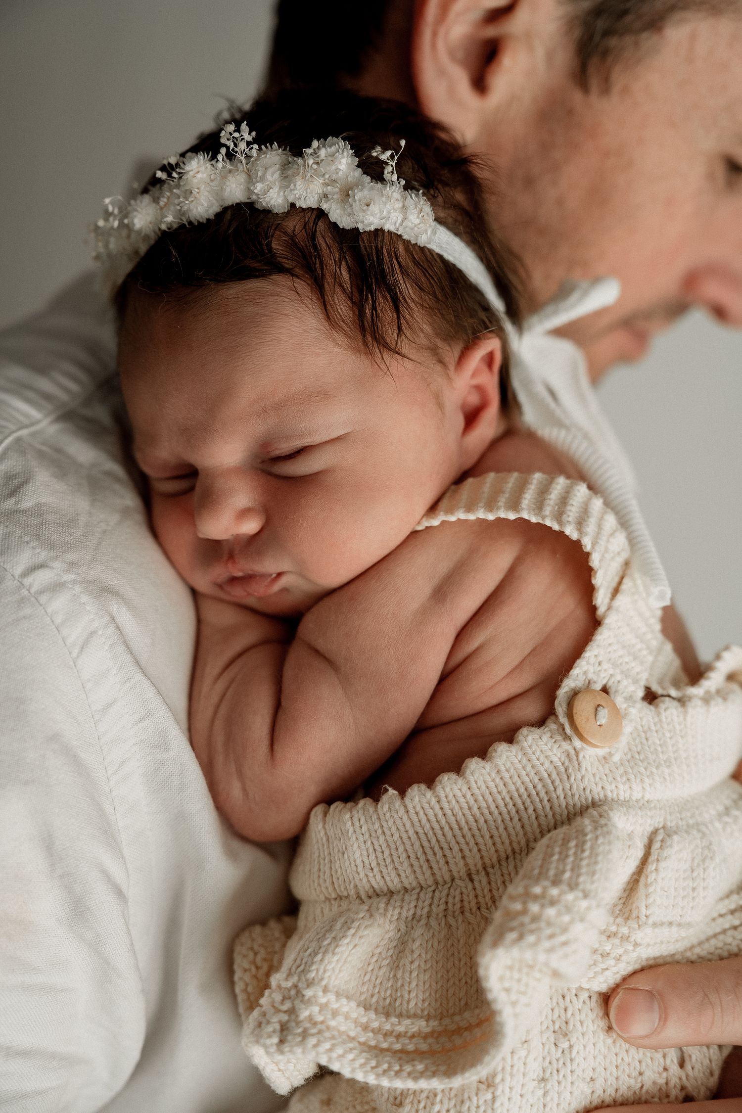 Peaceful sleeping newborn baby wearing white knit sweater and delicate lace headband on soft bedding.