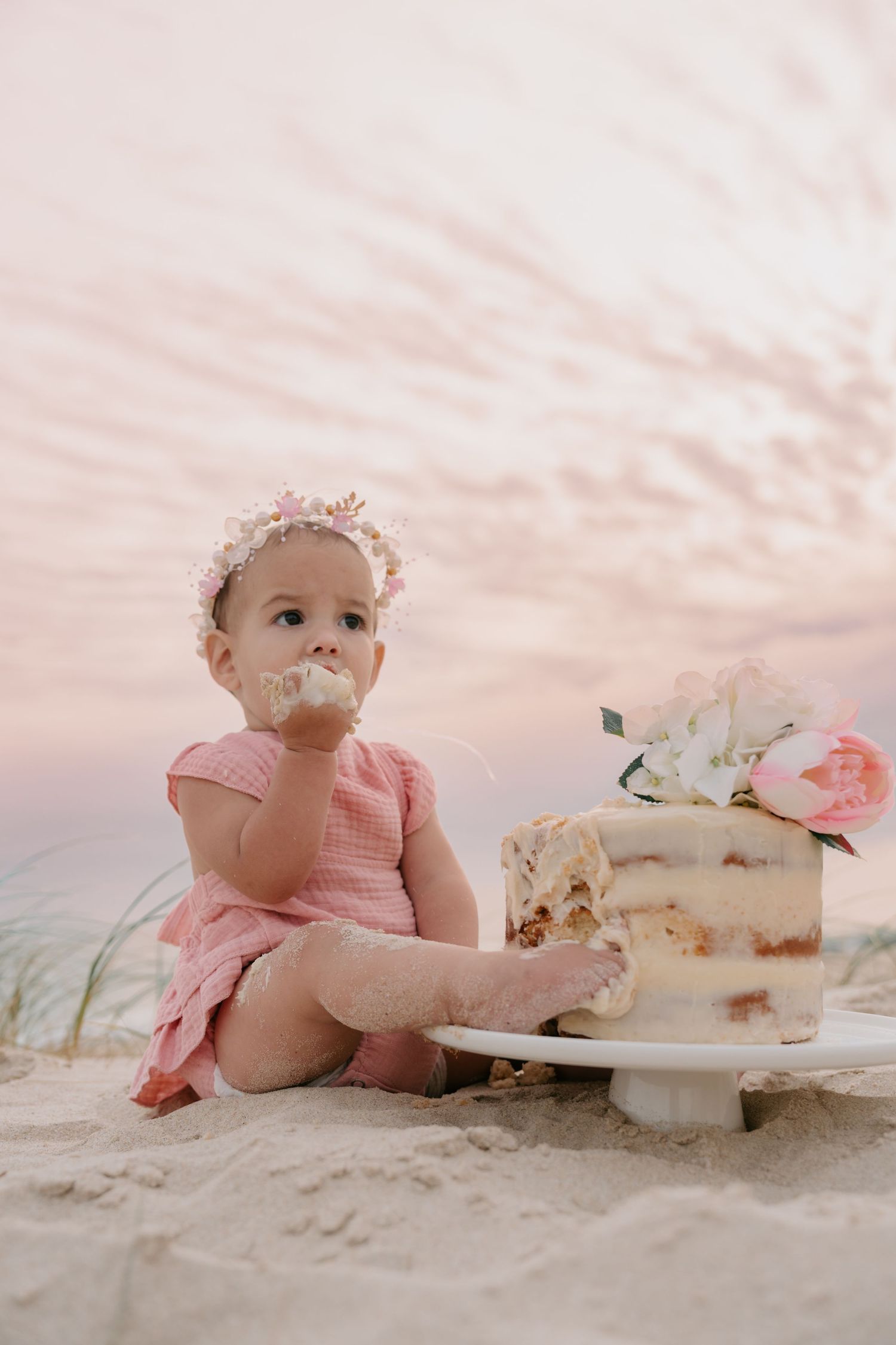 Baby girl in pink dress eating first birthday cake on sandy beach at sunset.