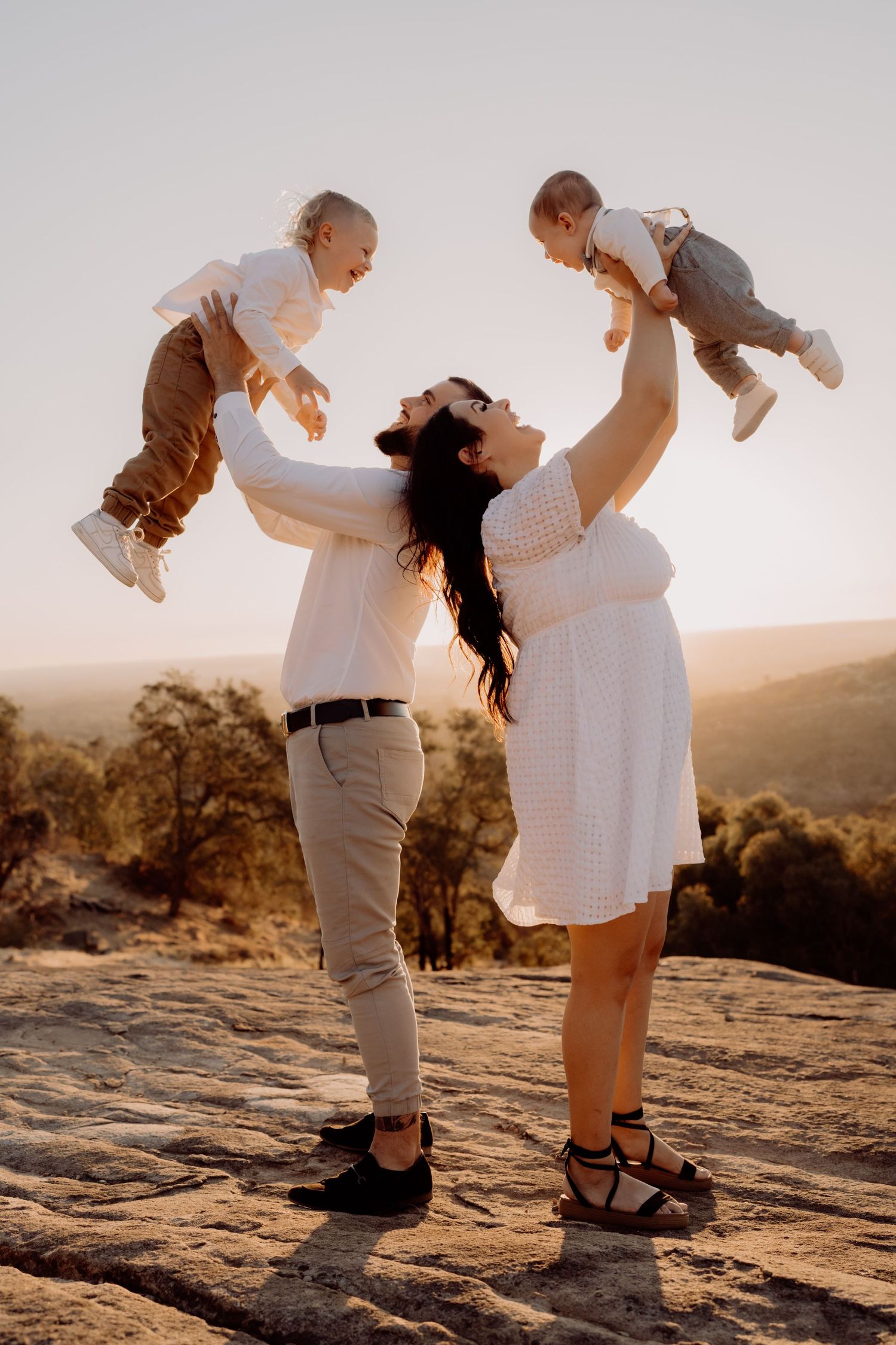 A couple in white outfits playfully lifts their child against a desert sunset backdrop at golden hour.