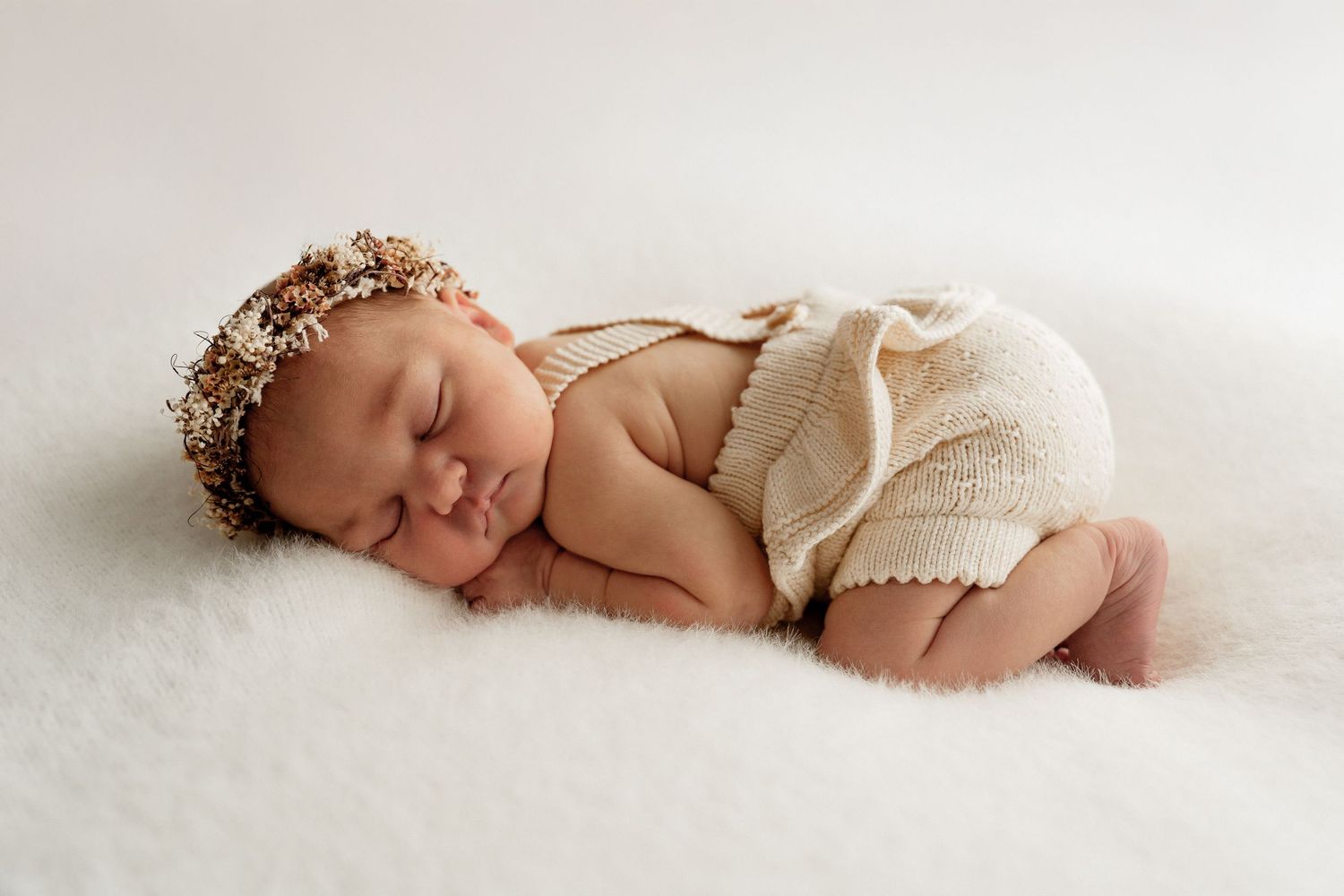 Sweet newborn in cream knit romper and delicate floral crown peacefully sleeping on soft white blanket.