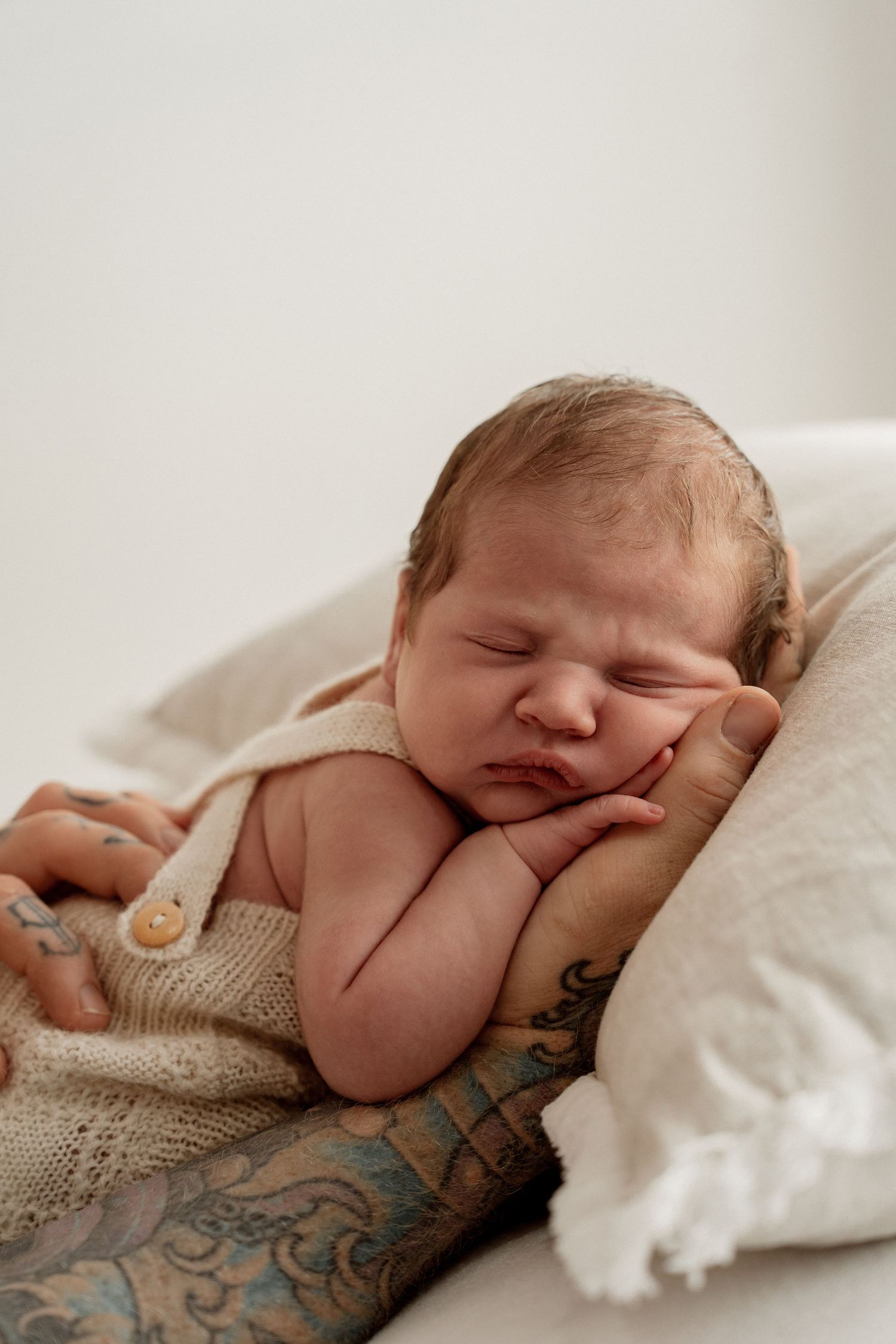 Sleeping newborn rests peacefully on tattooed arm while nestled in cozy beige knit blanket.