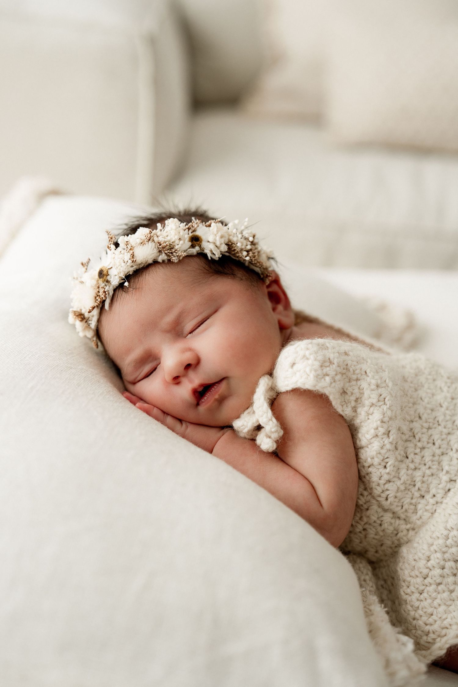 Sleeping newborn baby girl wearing a floral headband and cream knit outfit, peacefully resting on a white blanket.