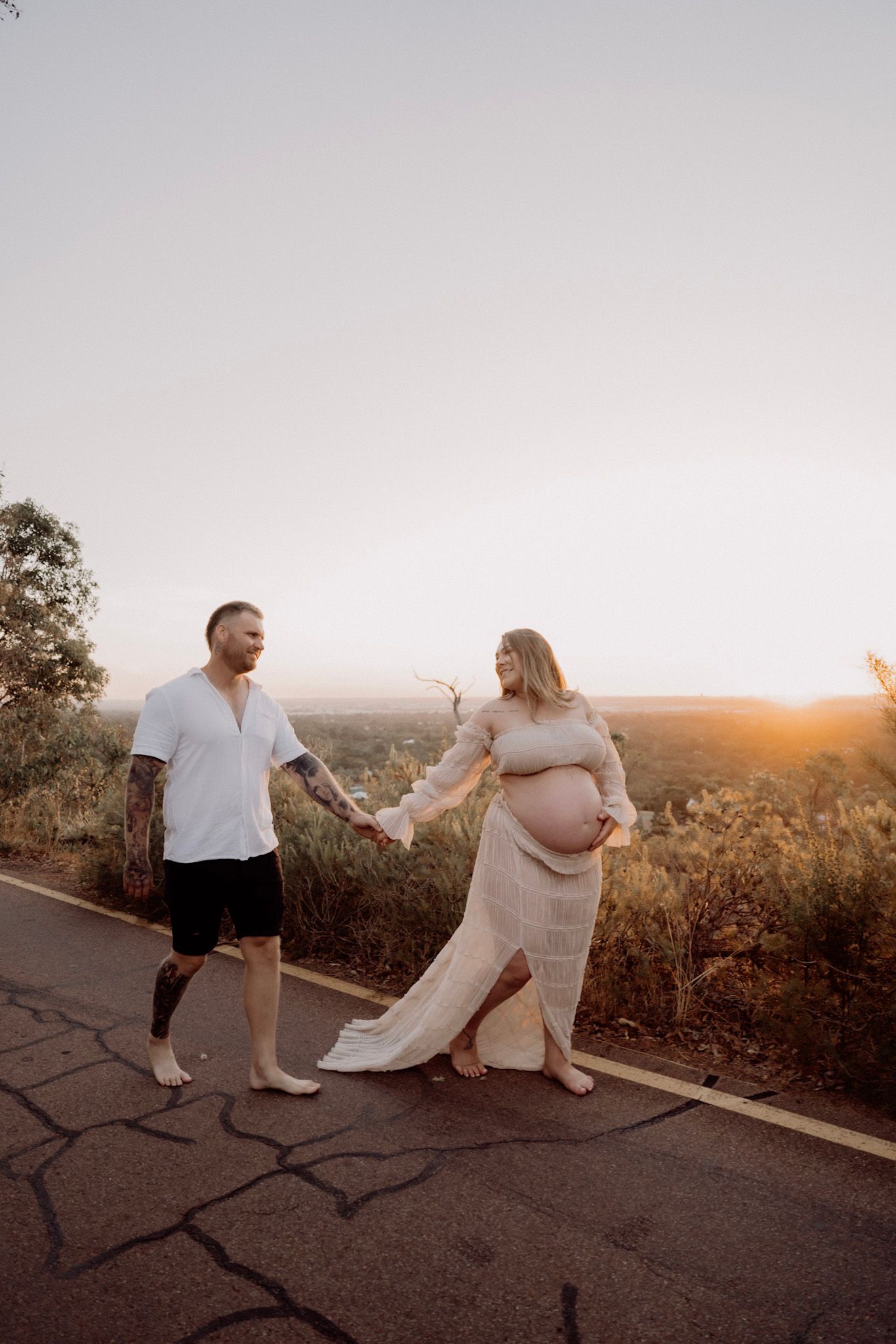 A couple walks hand in hand on a scenic road during sunset, with the expectant mother wearing a flowing beige dress.