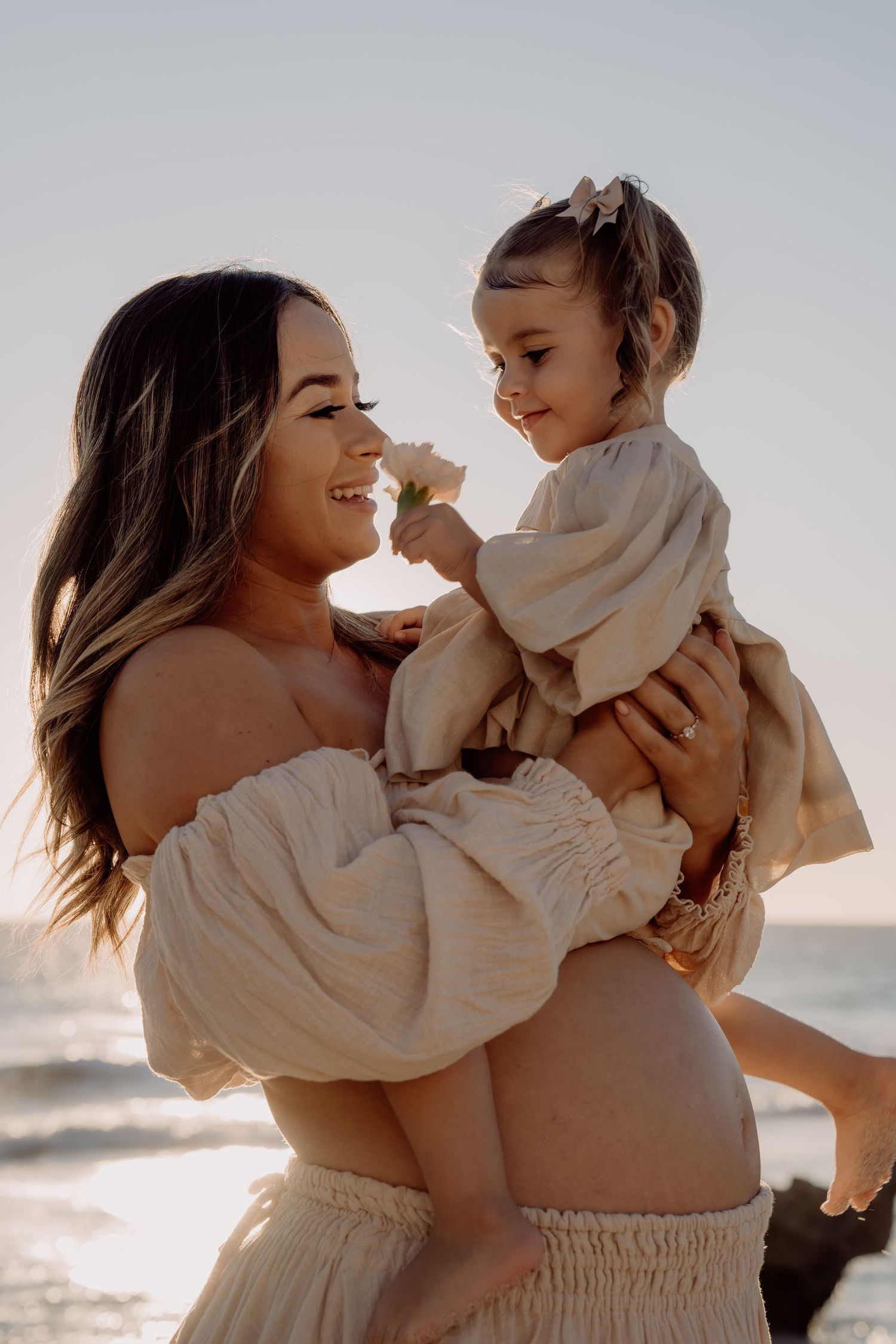 A parent and young child share a tender moment together on the beach at sunset, both wearing flowy white outfits.