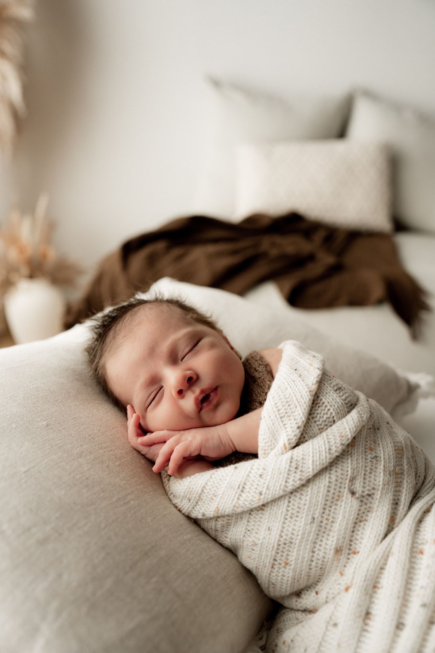 A peaceful newborn baby sleeping soundly, swaddled in a white knit blanket on a soft linen pillow.
