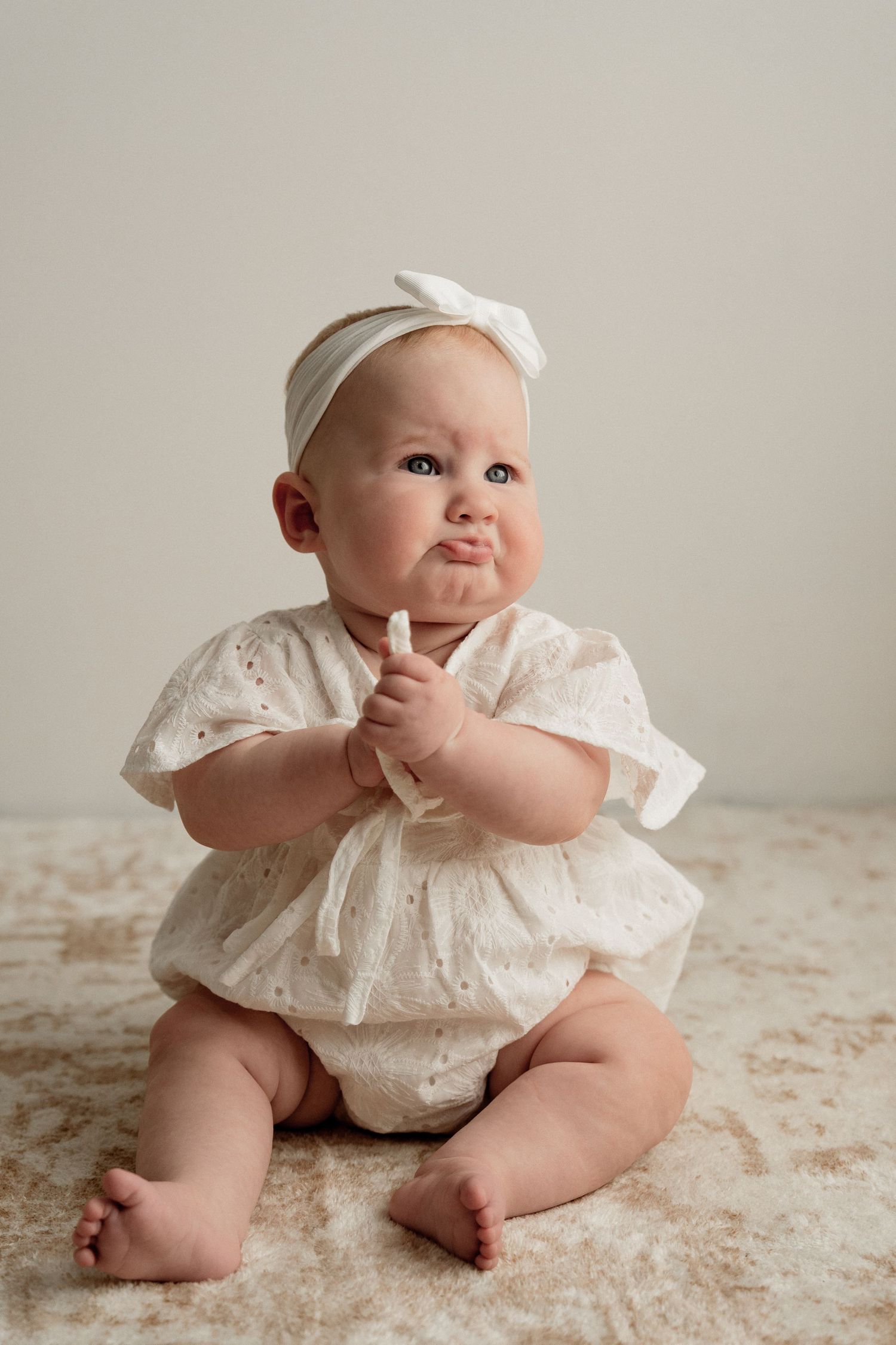 A baby wearing a white lace dress sits on a textured carpet in a series of sweet candid poses.
