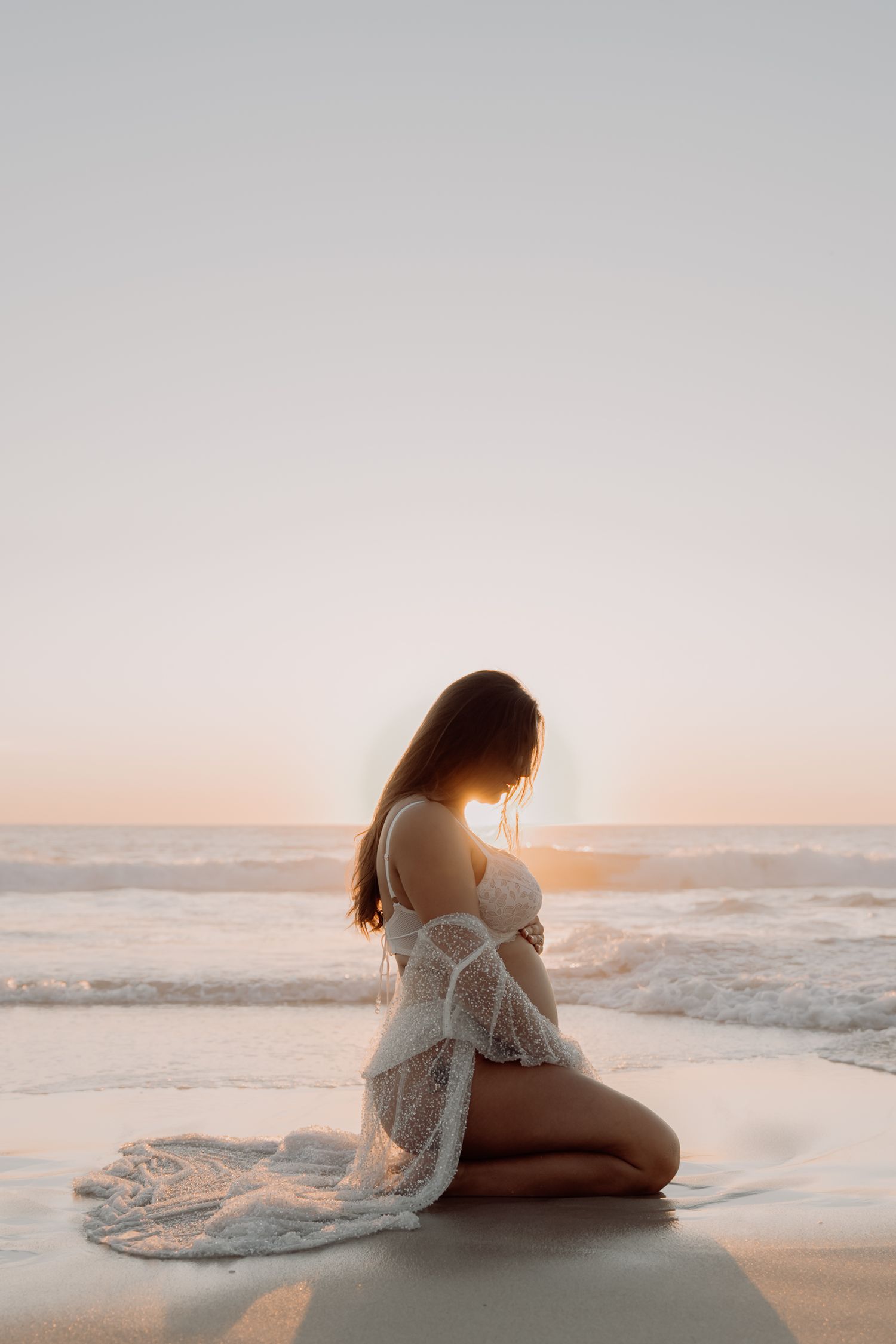 A person in a white lace dress sits on the beach during golden hour sunset with waves crashing in the background.