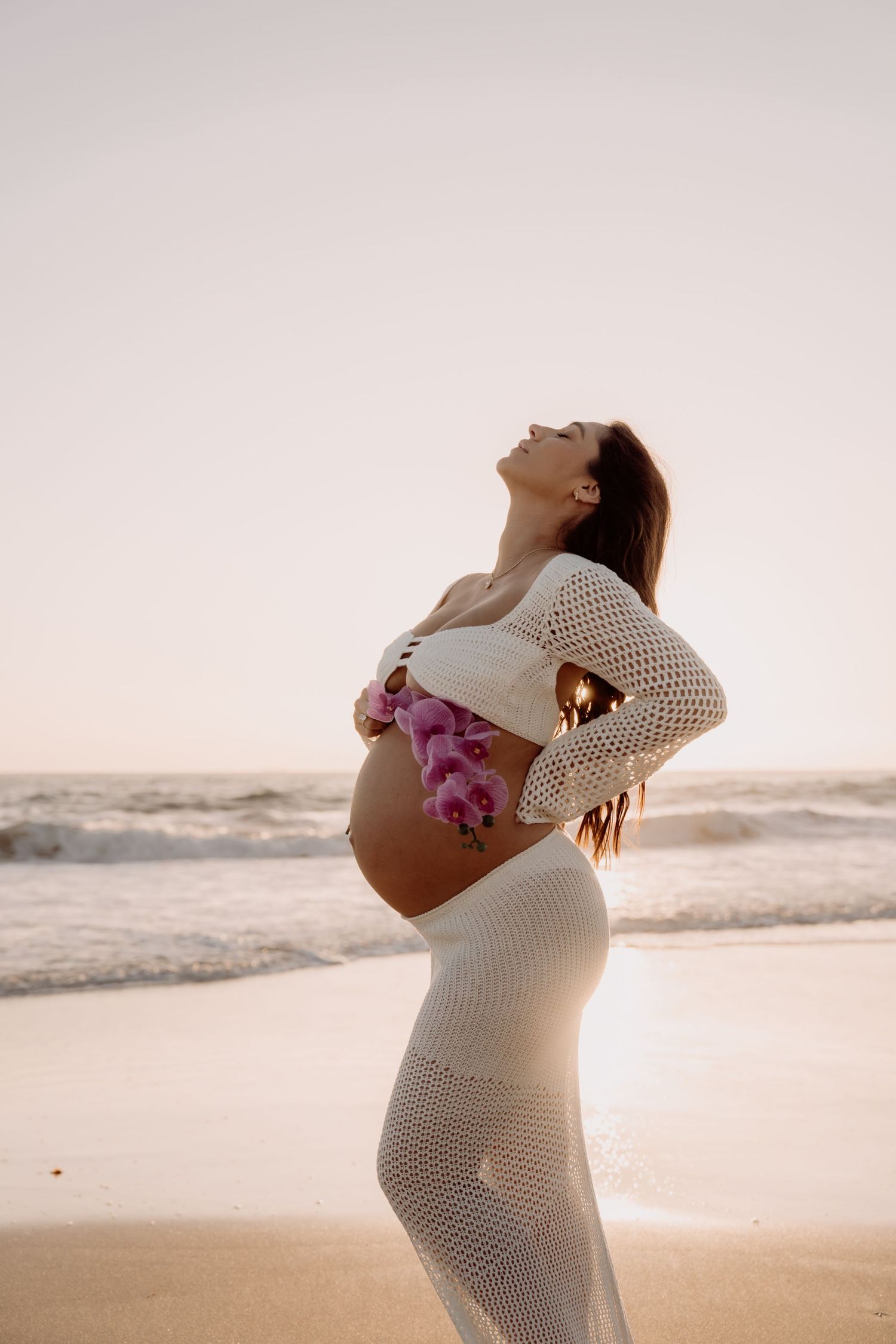 Silhouetted pregnant figure in crochet dress stands on beach at sunset, arching back in serene moment.