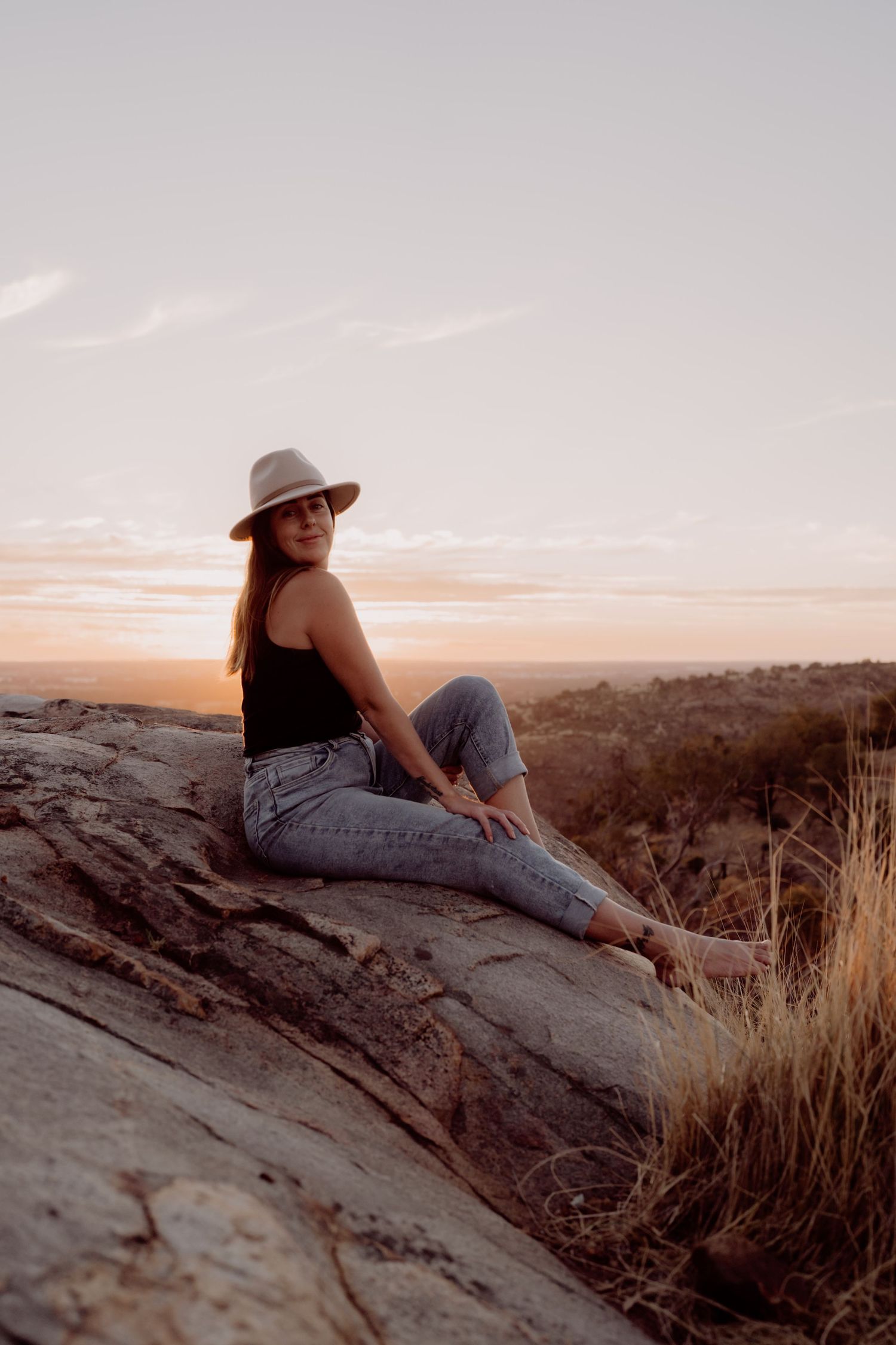 Woman in hat and jeans sitting on rock at golden hour sunset with vast landscape in background.