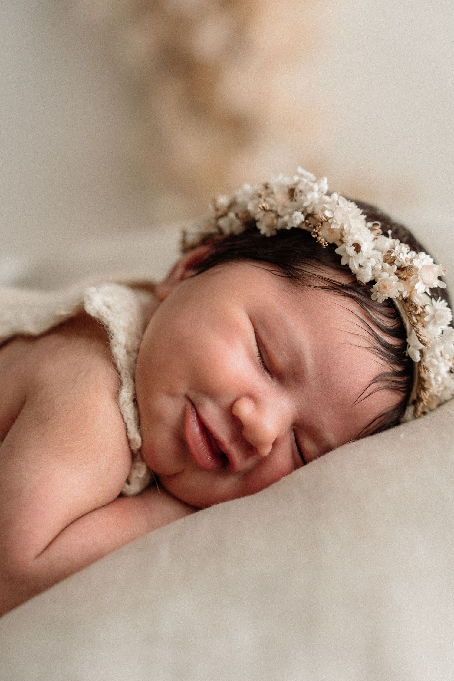 Sleeping newborn wearing delicate white floral crown rests peacefully on soft beige blanket.