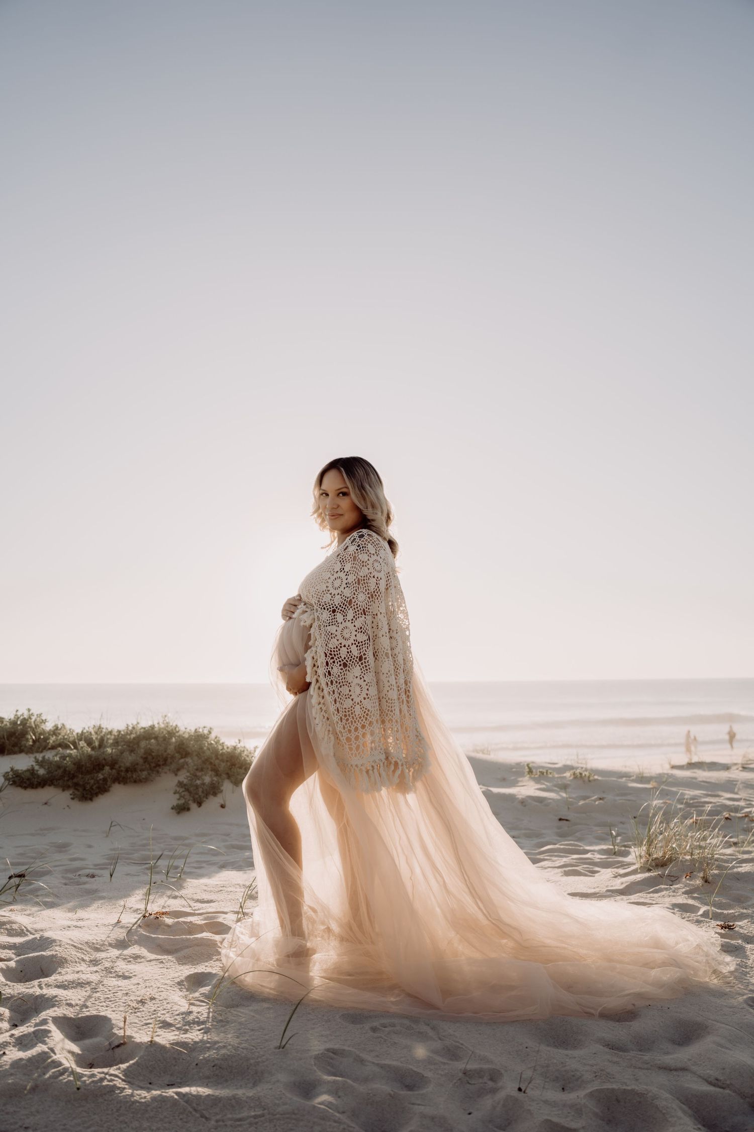 A flowing lace wedding dress elegantly trails across sandy beach dunes at sunset, creating a romantic coastal scene.