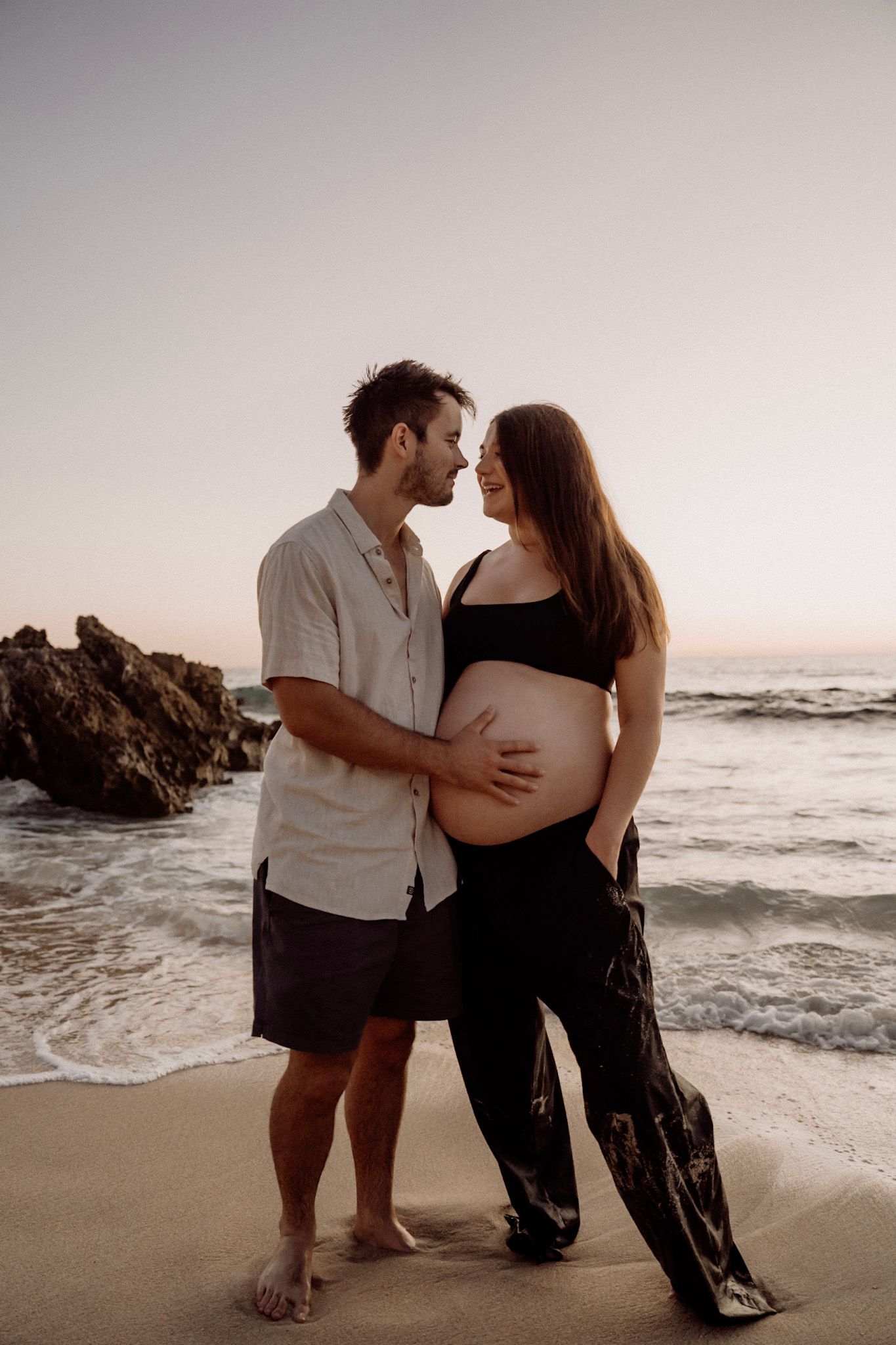 Romantic couple shares intimate moments at sunset on a rocky beach overlooking ocean waves.
