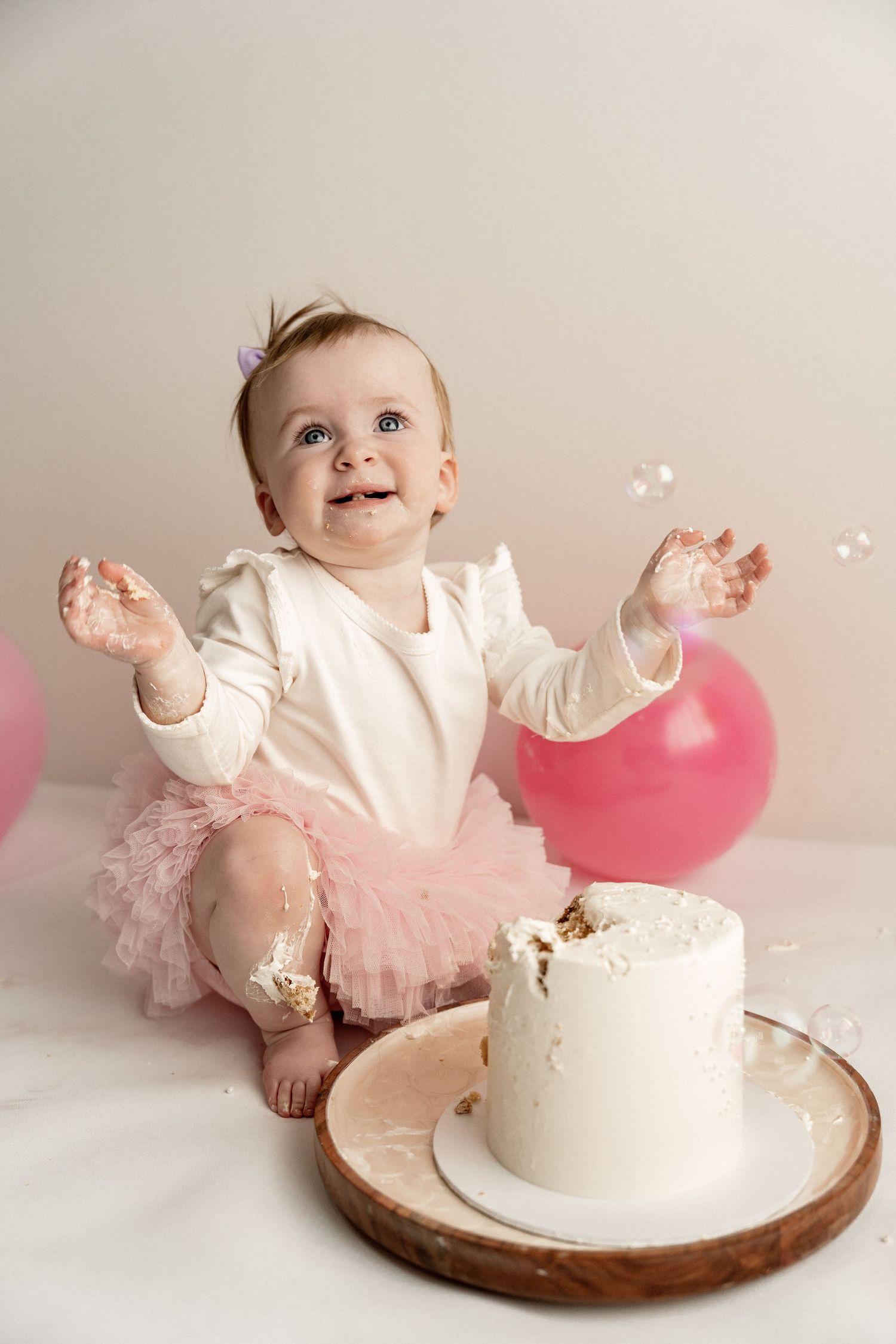 Baby in pink tutu and white top smashes into white birthday cake during first birthday cake smash photo session.