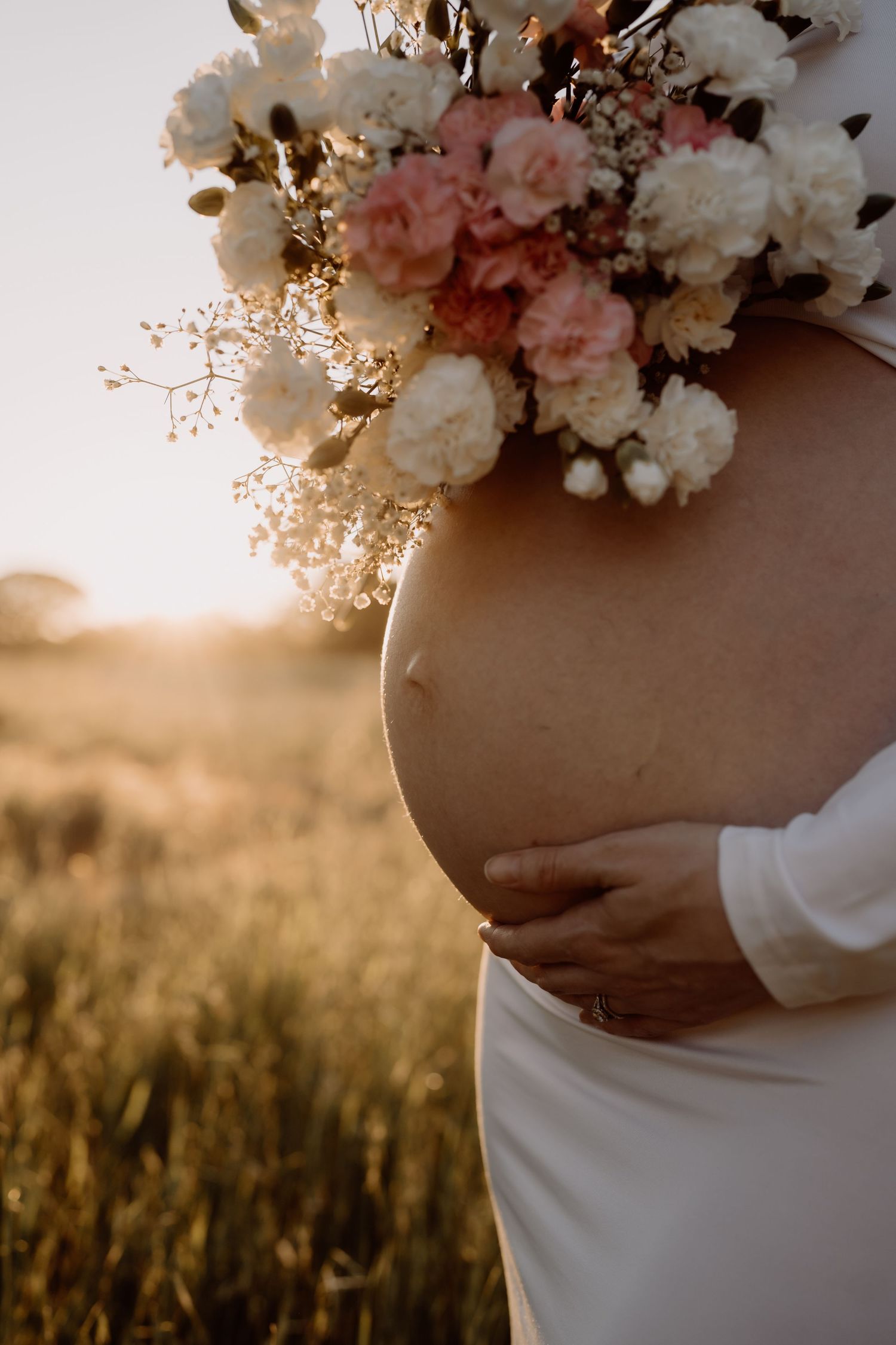Delicate pink and white floral bouquet held against a pregnant belly in white dress at golden sunset in field.