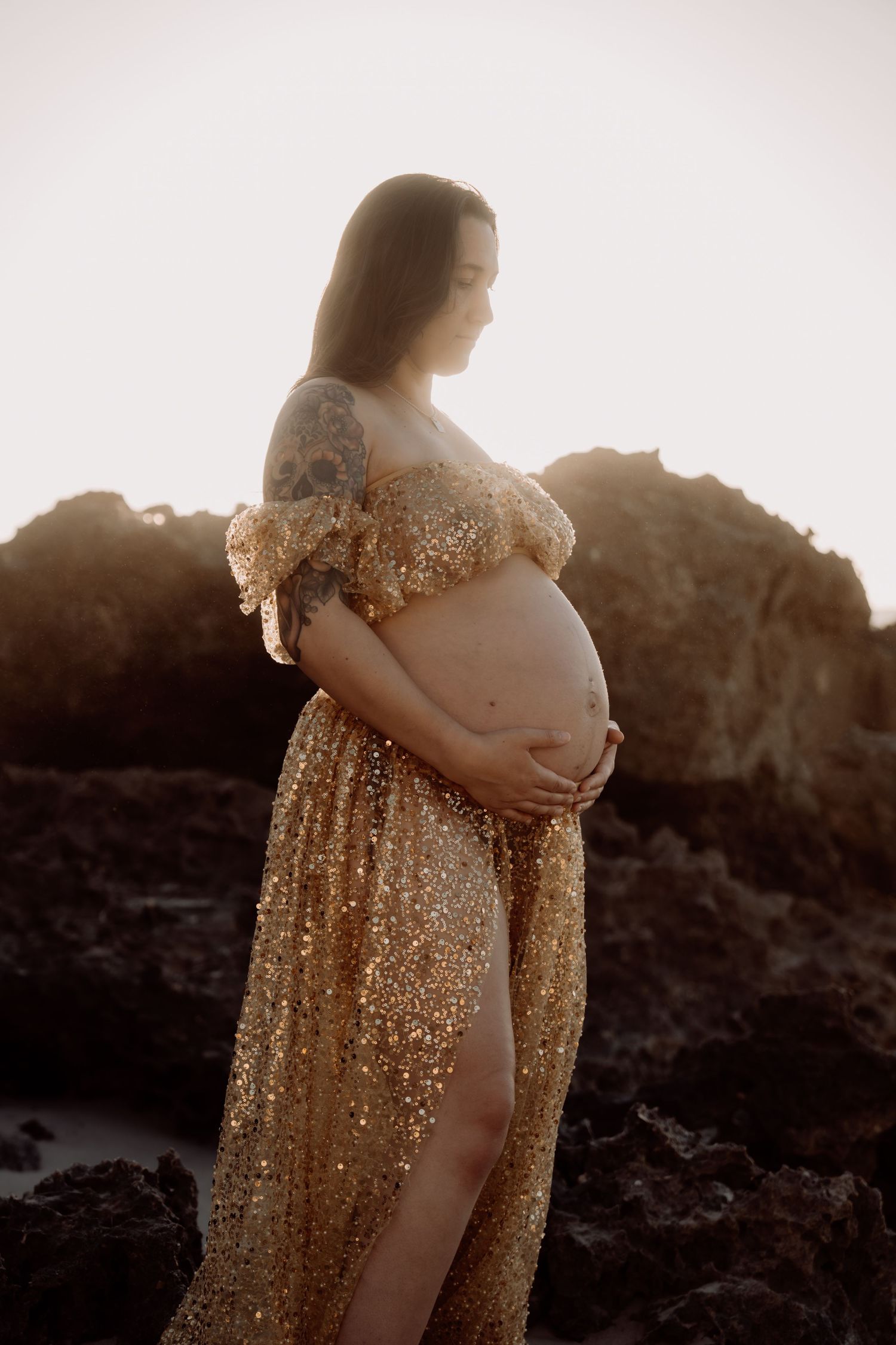 A silhouetted figure in a sparkling gold maternity gown poses against a dramatic sunset backdrop during a pregnancy photoshoot.