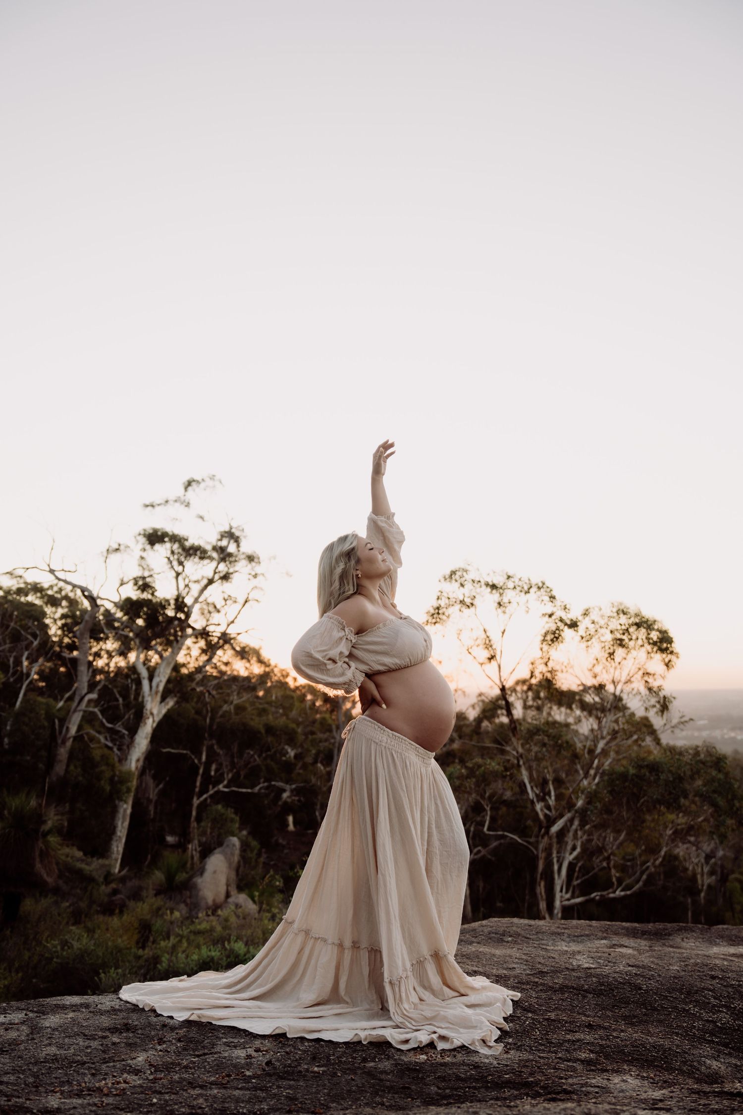 Pregnant woman in beige flowing gown poses gracefully on rocky outcrop at sunset with eucalyptus trees behind her.