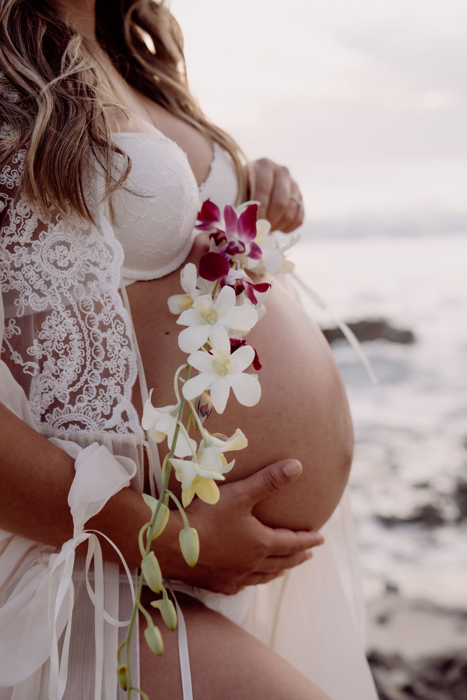 Pregnant woman in white lace robe holding baby bump with cascading orchid flowers near ocean shore.
