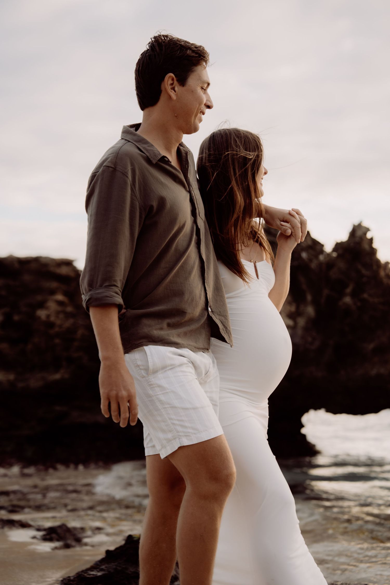 Couple poses on rocky beach, pregnant woman in white dress beside man in gray shirt and white shorts.