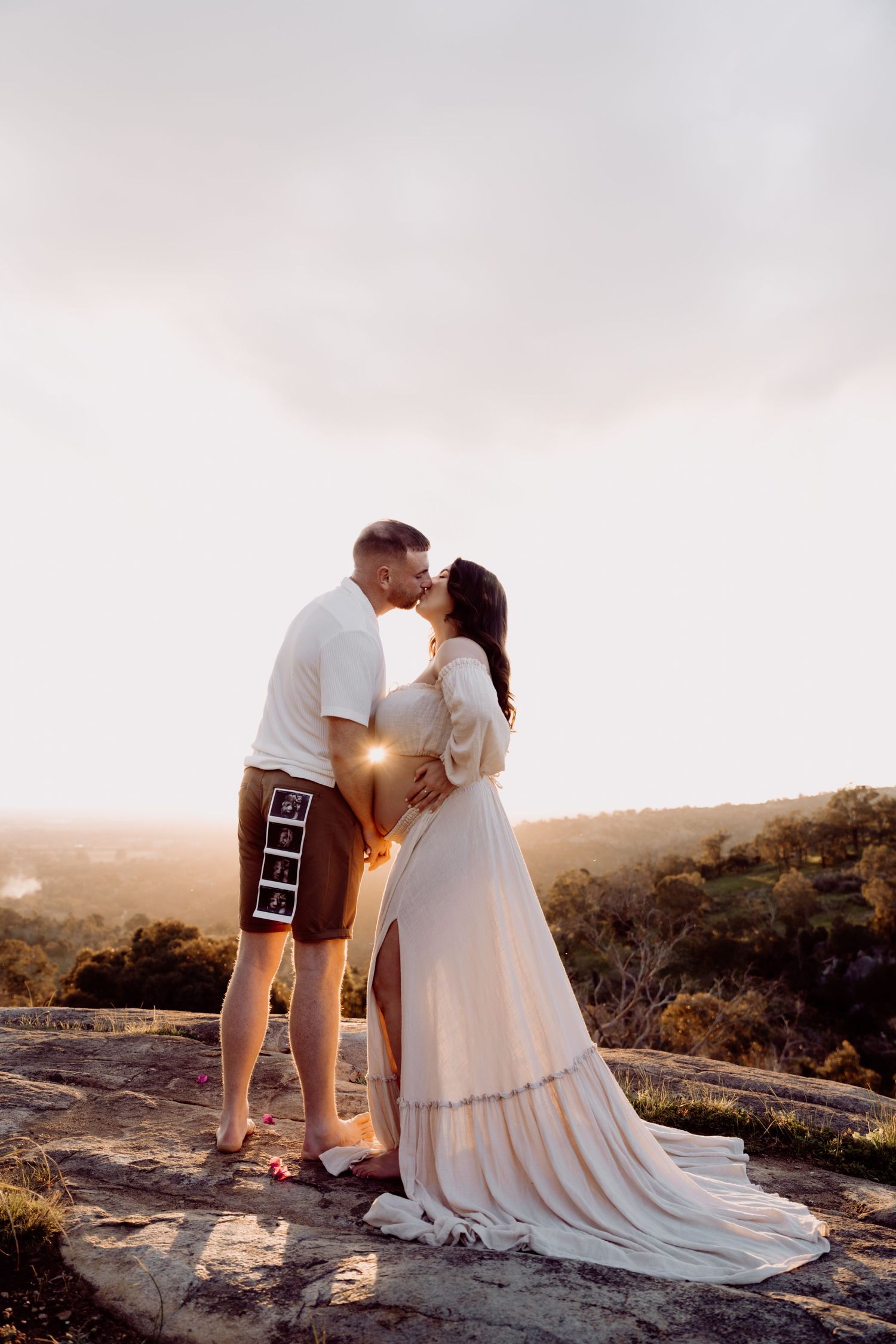 Couple sharing a kiss at golden hour on a rocky hilltop, woman in flowing white maternity dress holding ultrasound photos.
