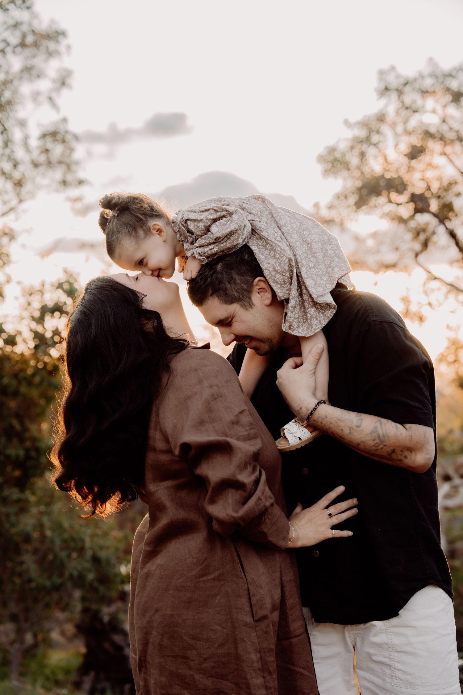 A couple sharing a tender kiss while holding their young child outdoors during golden hour sunset.
